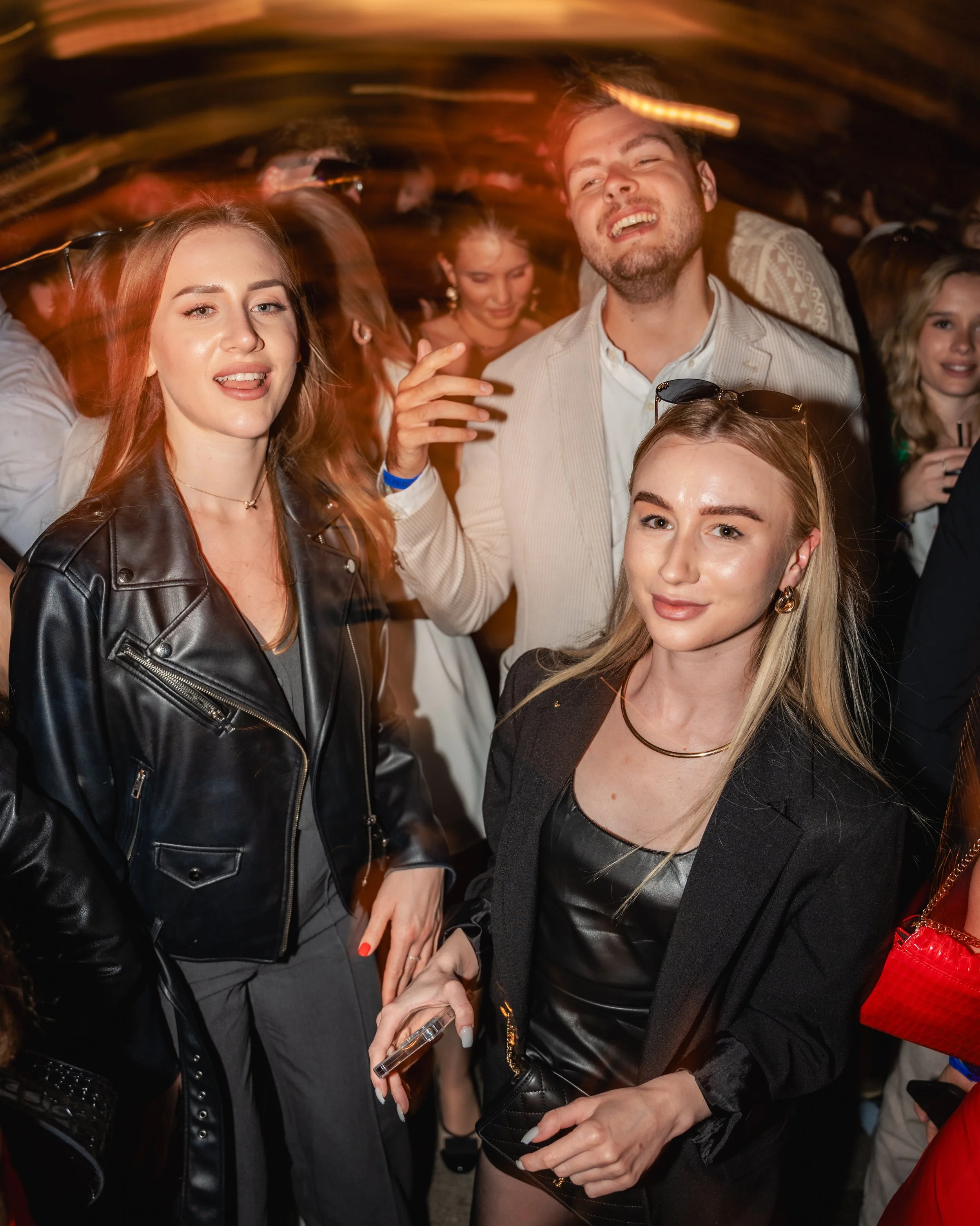 A group of young adults socializing at a party or nightclub, with a focus on two women in the foreground dressed in black leather and a man in a light-colored blazer behind them, all smiling or enjoying the moment.