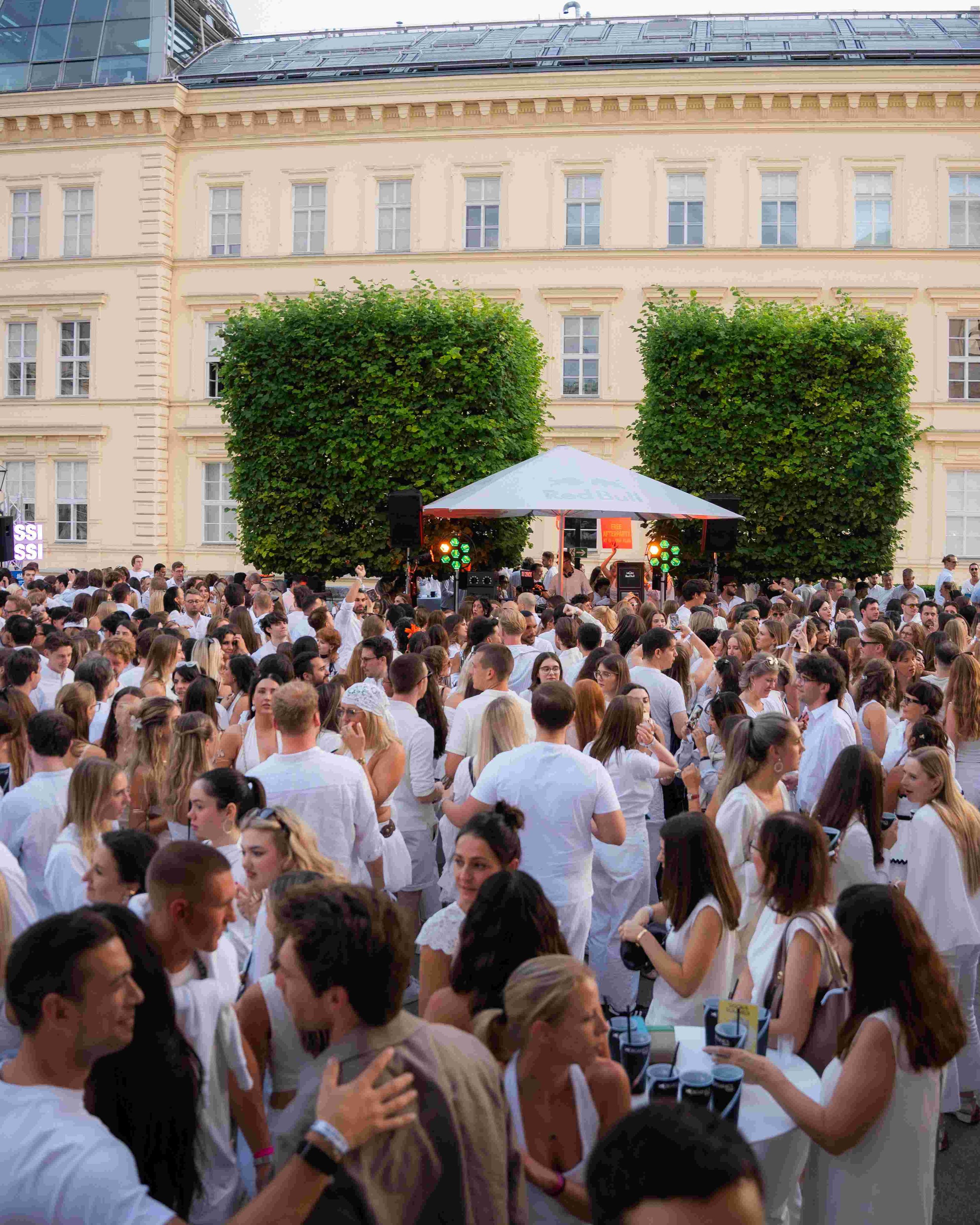 Crowd of people wearing white clothing at an outdoor event with a stage and large building in the background, surrounded by green trees.