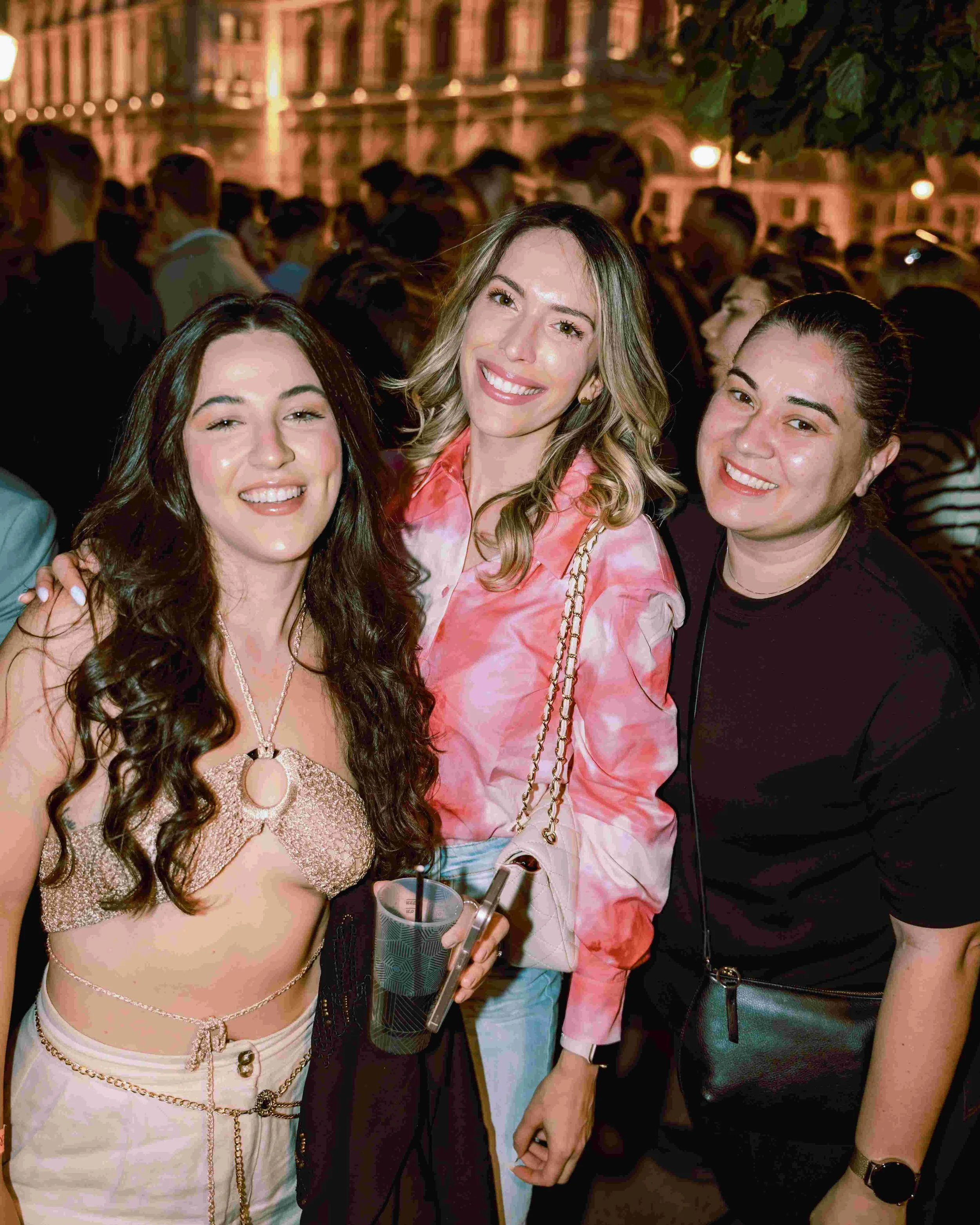 Three women standing close together at a nighttime outdoor event, smiling at the camera, with a large crowd in the background.