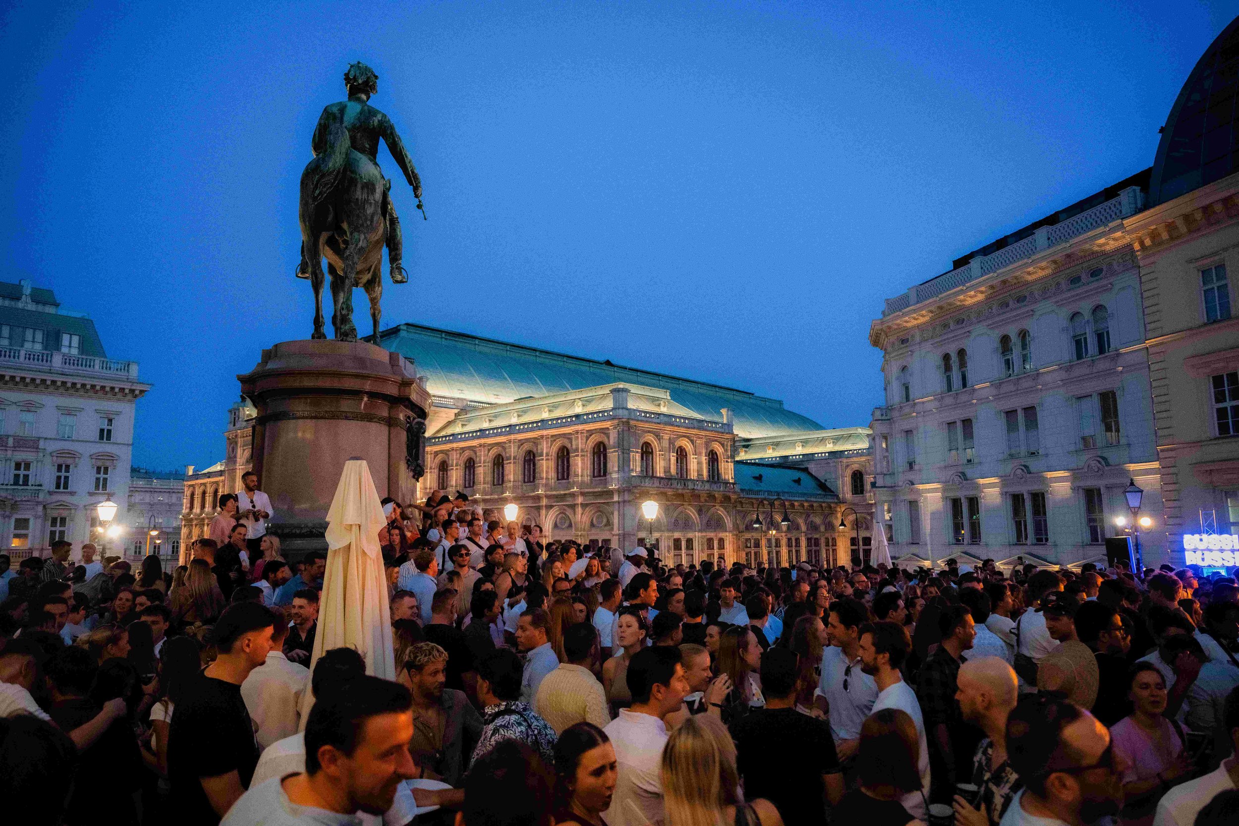Crowd of people gathered in an outdoor city square during evening, with historic buildings and a statue of a man on a horse in the background.