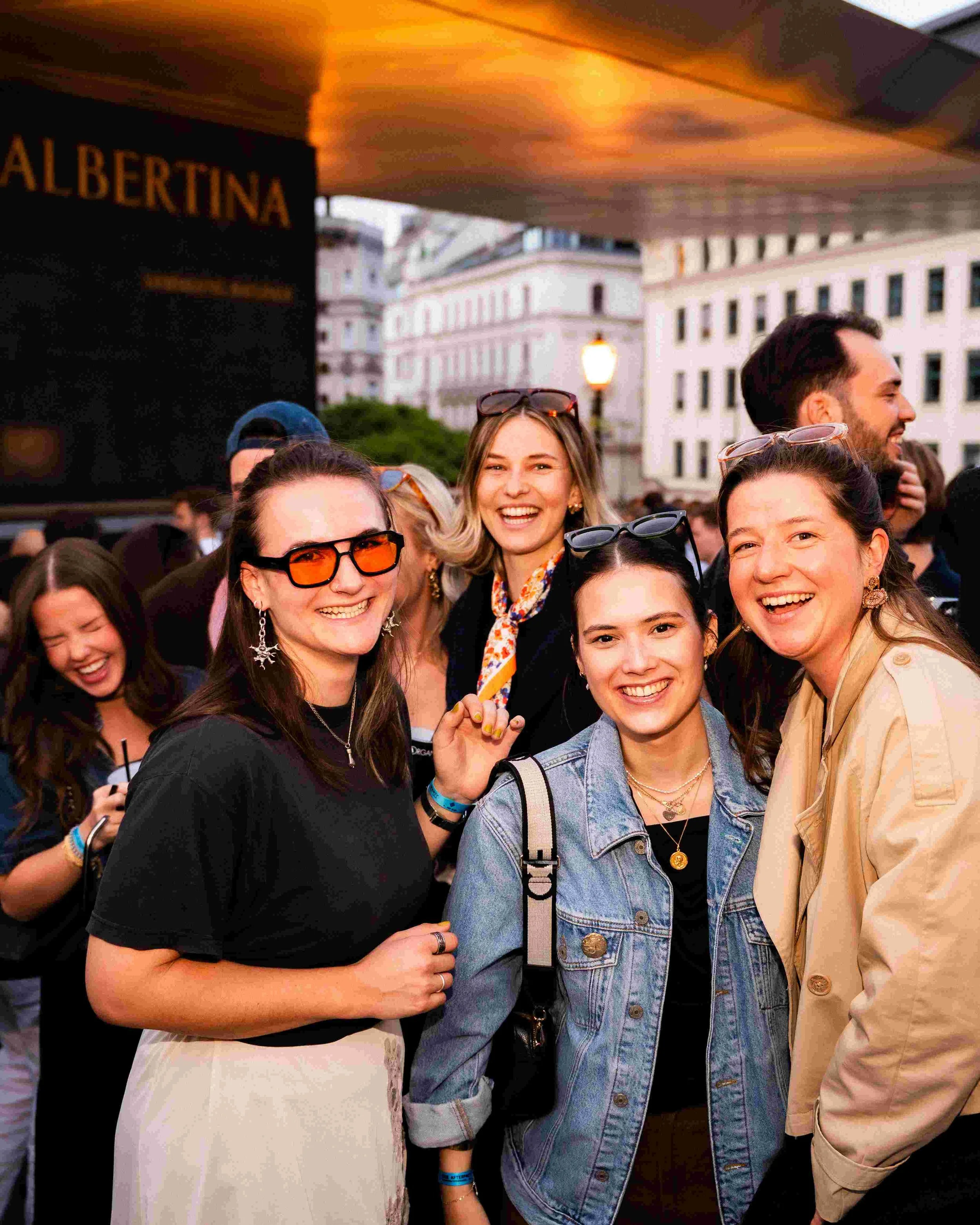 Group of smiling young women and young men gathered outdoors during sunset, with historic buildings in the background.