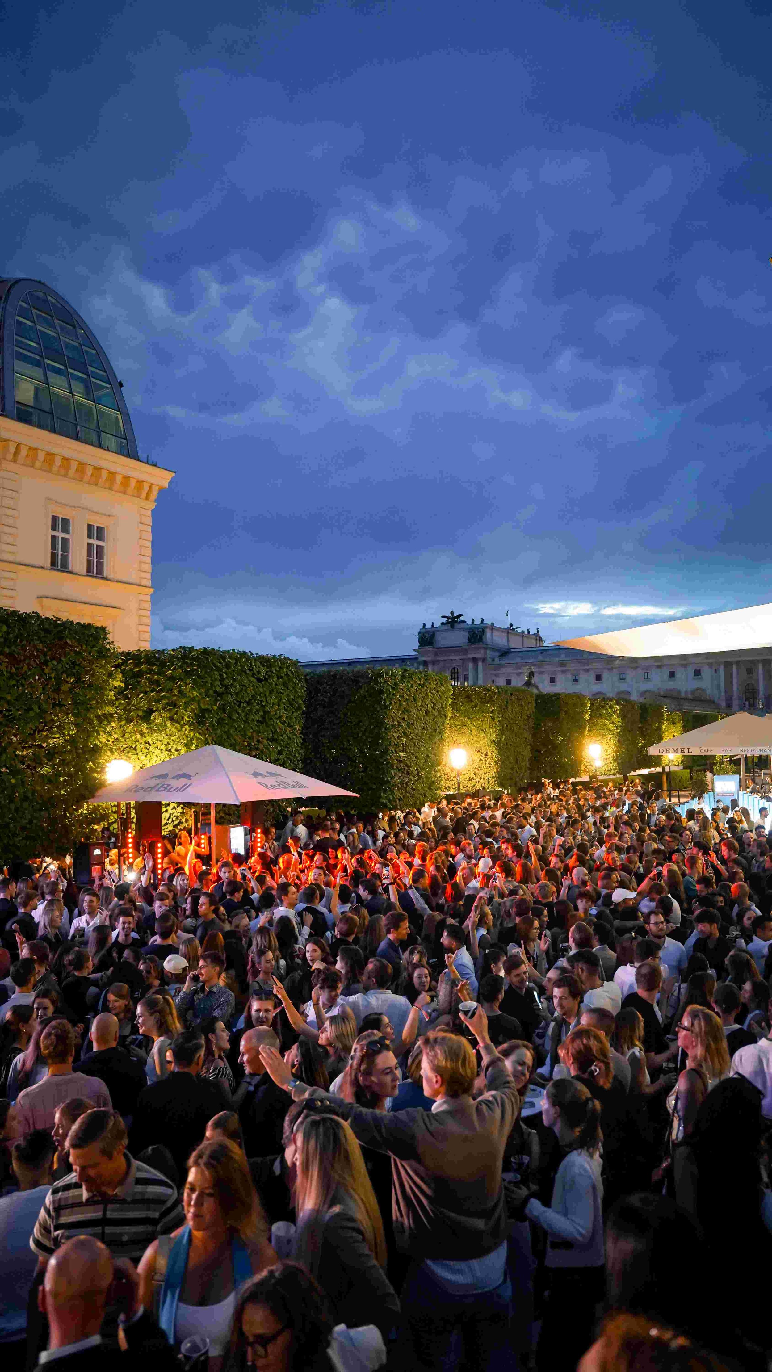A large crowd of people gathered outdoors during the evening, with some holding drinks and engaging in conversations. The scene is illuminated by street lamps and a tent with a Red Bull logo, under a cloudy sky.