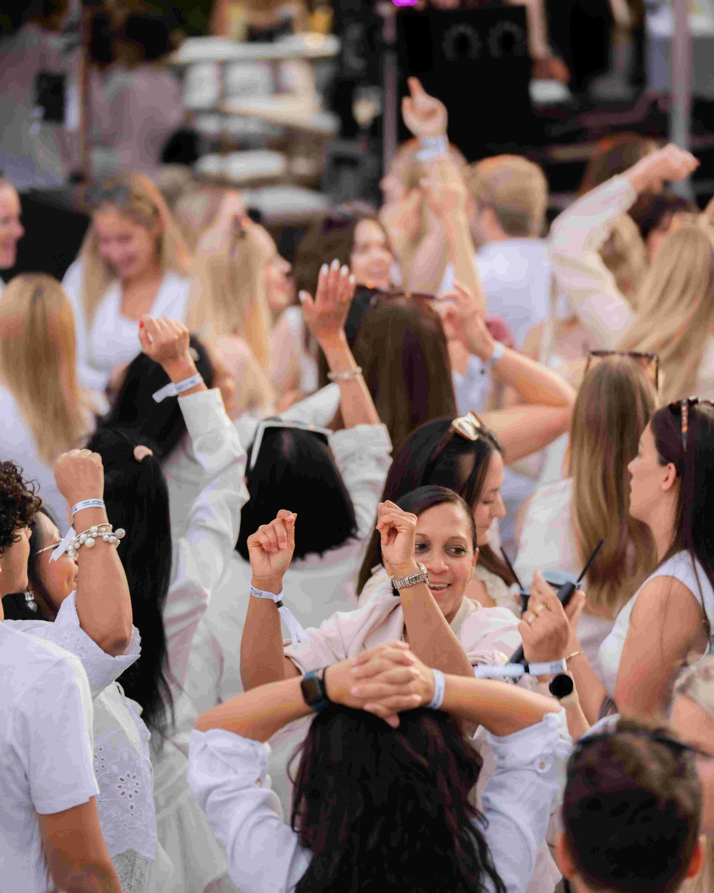 A group of women wearing white clothing, gathered close together, appearing to be at a social event or celebration, with some smiling and raising their hands.
