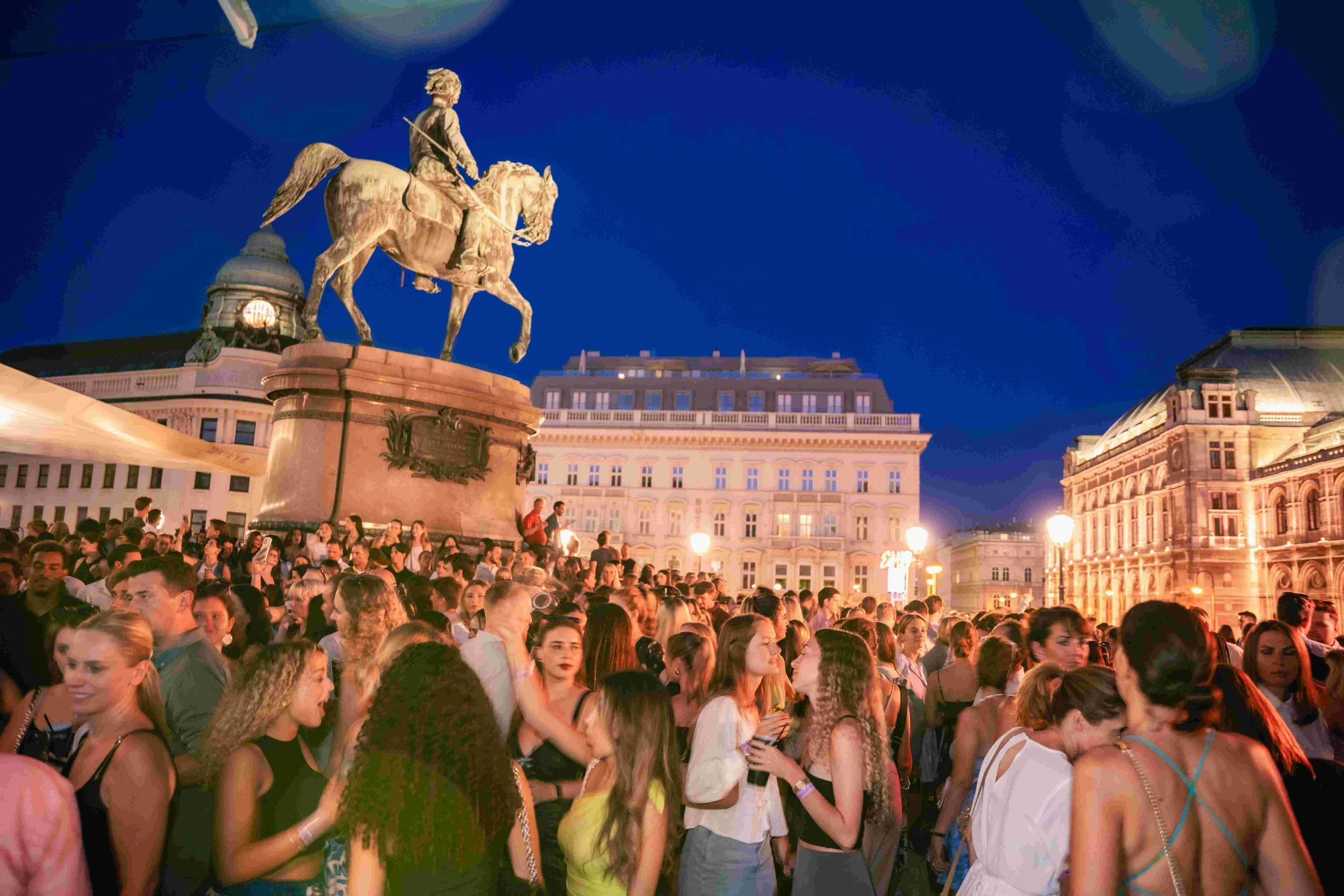 Crowd of people gathered outdoors at night in a city square with historic European-style buildings and a large equestrian statue.