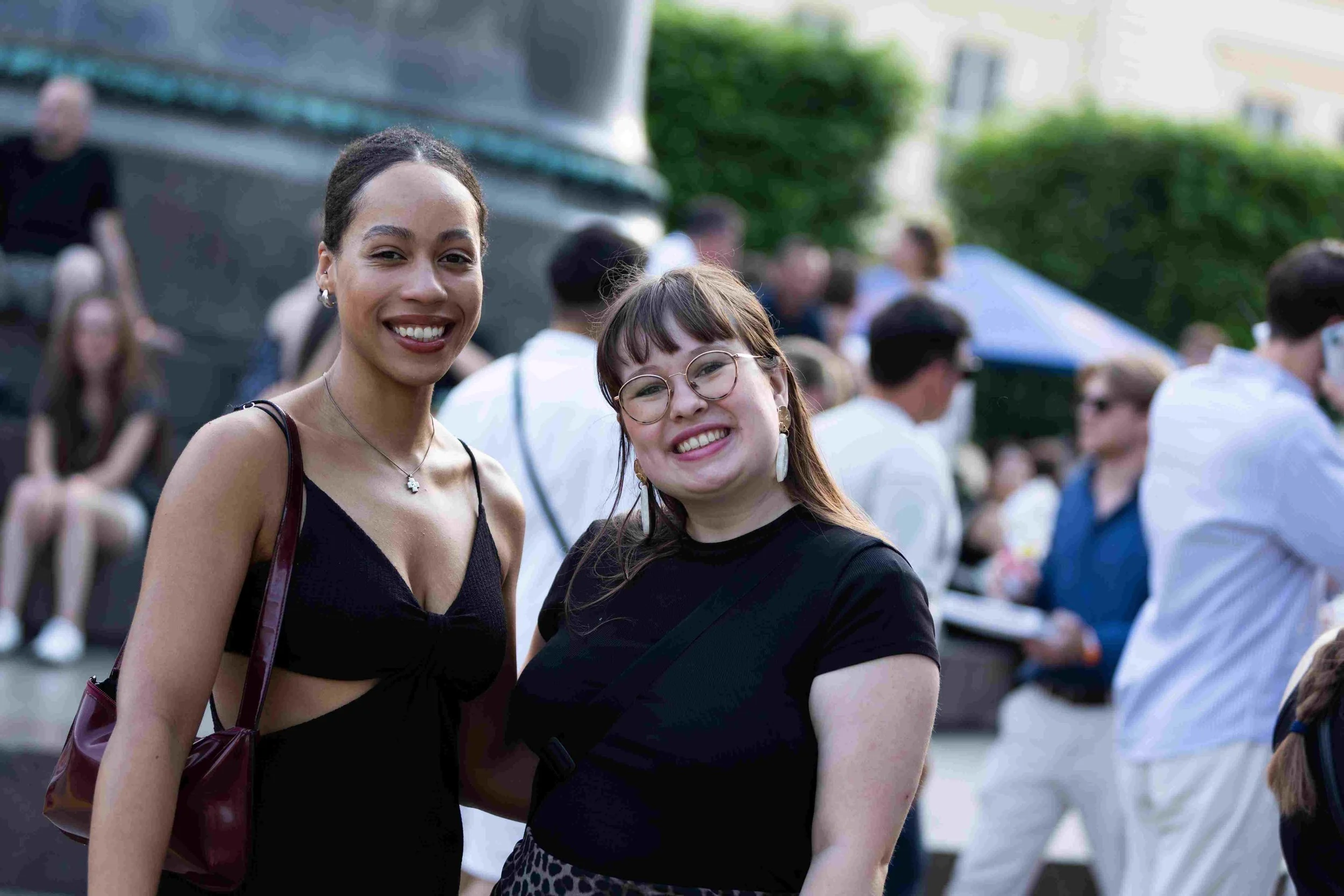 Two smiling young women standing outdoors at a crowded event, with other people in the background.