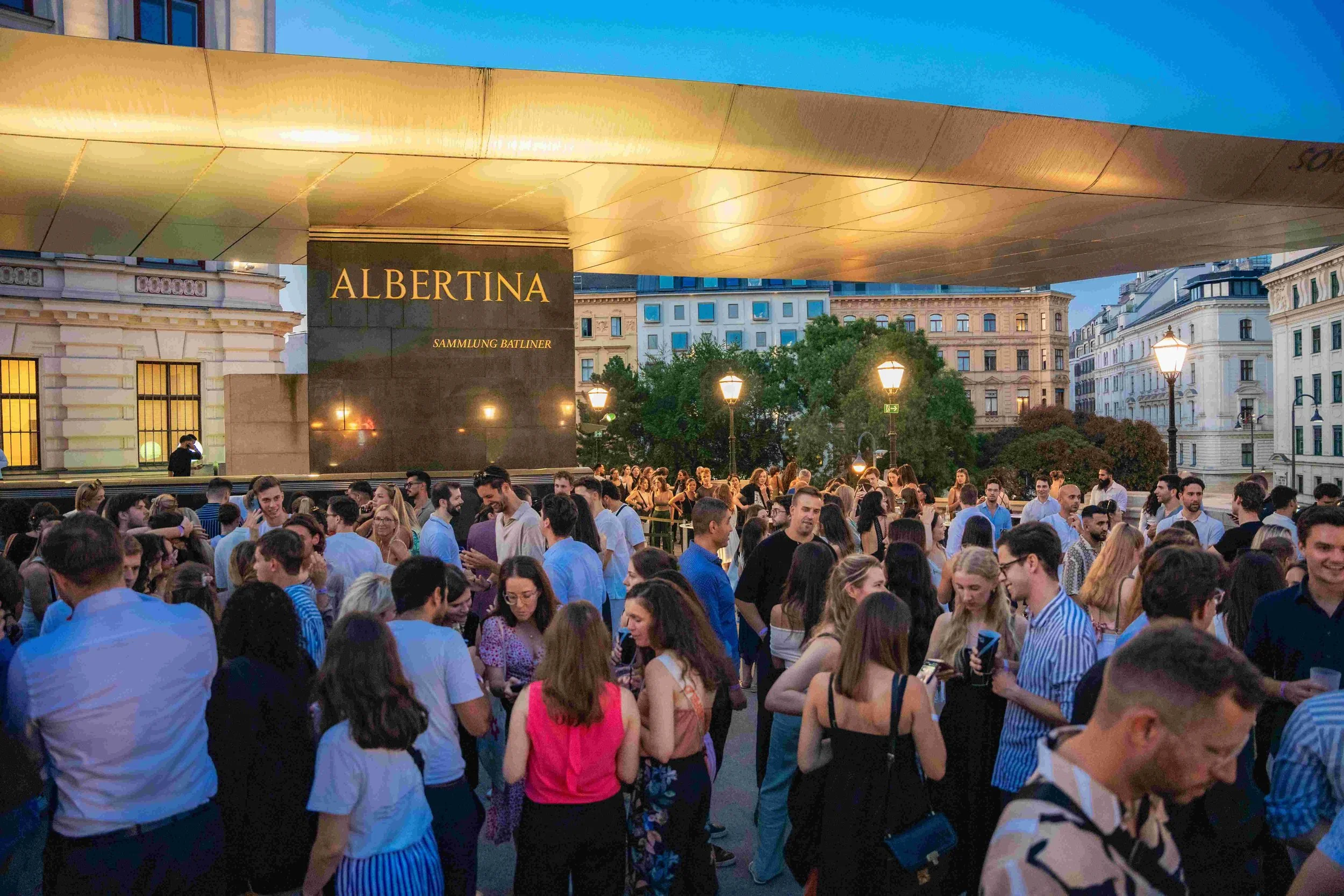 A crowd of people gathered outdoors at dusk under a large golden canopy with a sign reading 'ALBERTINA Sammlung Batliner.' Surrounding buildings and trees are visible in the background, illuminated by streetlights.