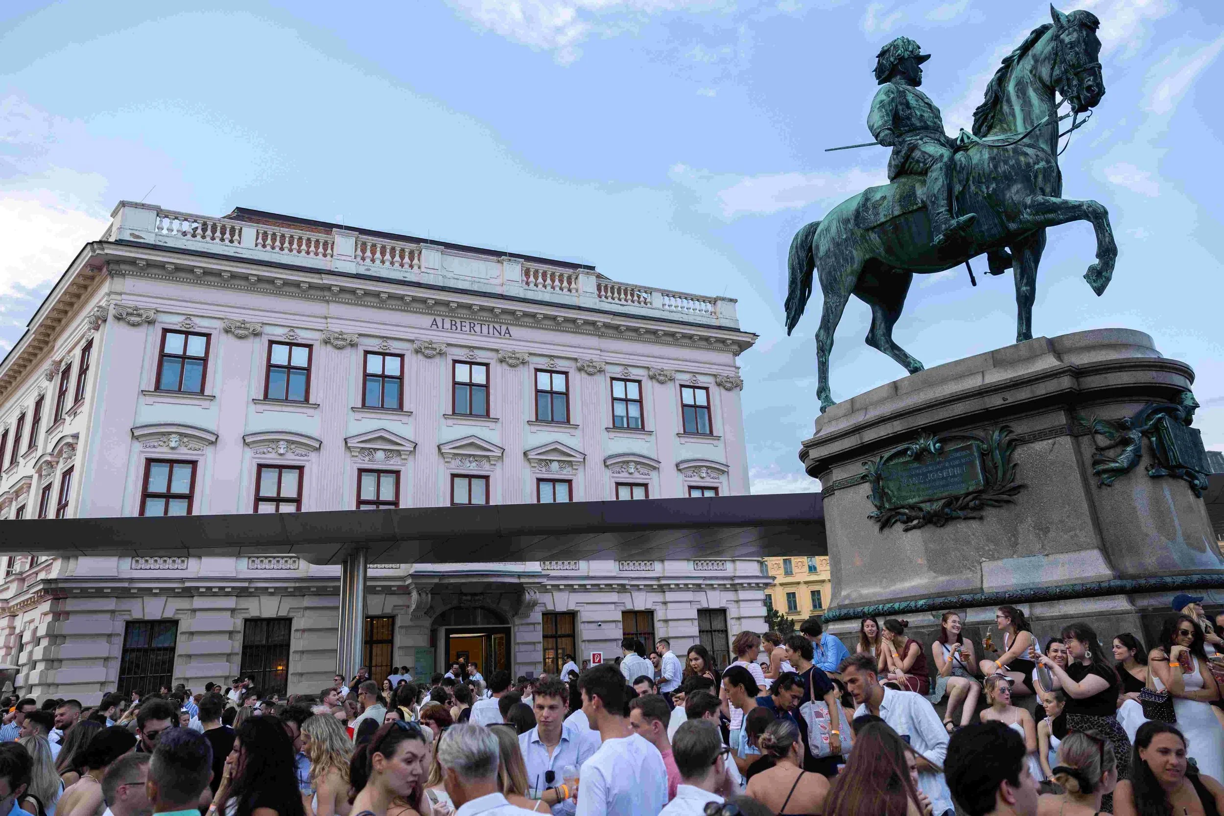 Crowd of people gathered in front of a historic building with a statue of a man on horseback. The building has the word 'ALBERTINA' on it, and the sky above is partly cloudy.