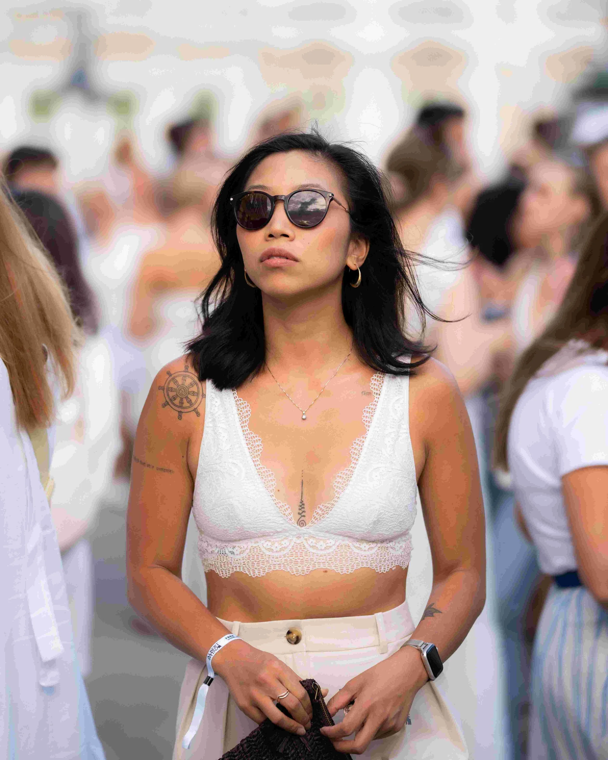 A woman with black hair, wearing sunglasses, a white lace tank top, gold hoop earrings, a necklace, a smartwatch, and a wristband, stands in a crowd.