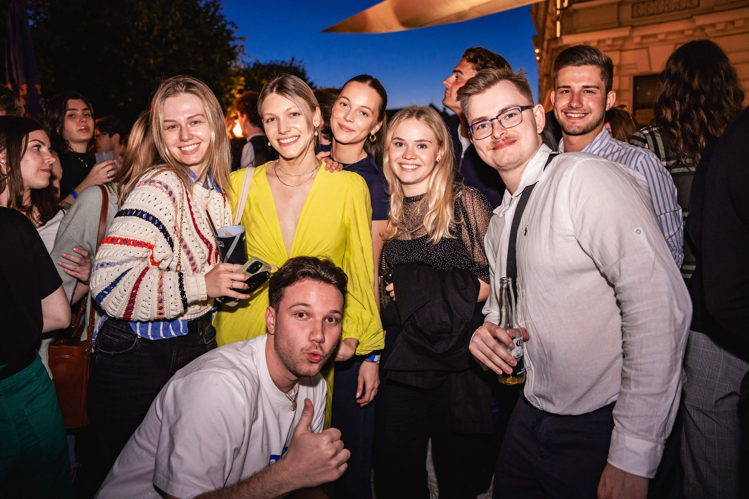 Group of young adults at an outdoor evening social event, smiling and posing for a group photo, with some holding drinks.