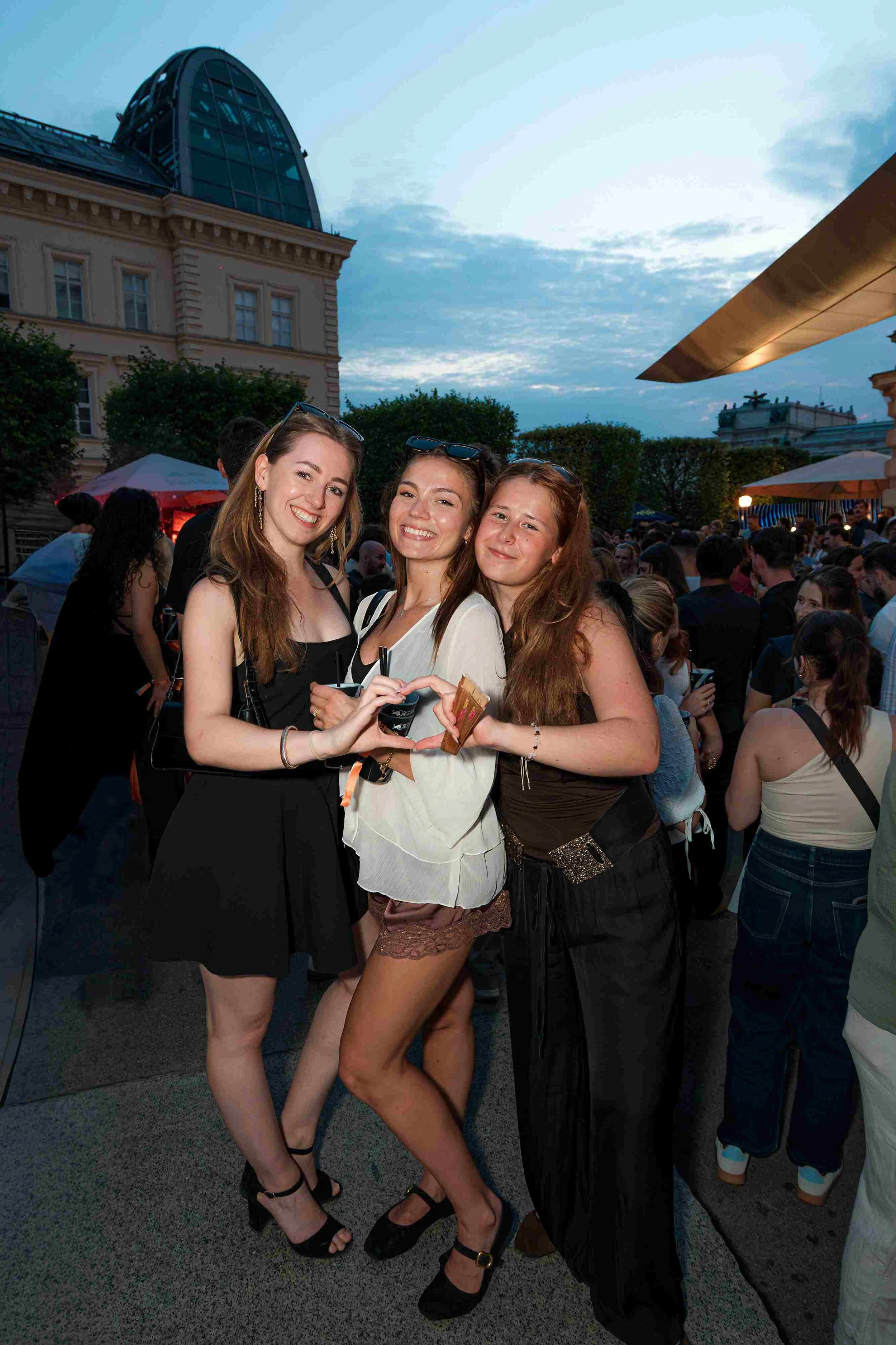 Three young women smiling and making a heart shape with their hands at an outdoor event during the evening, with a crowd behind them and a cityscape with historic and modern buildings in the background.