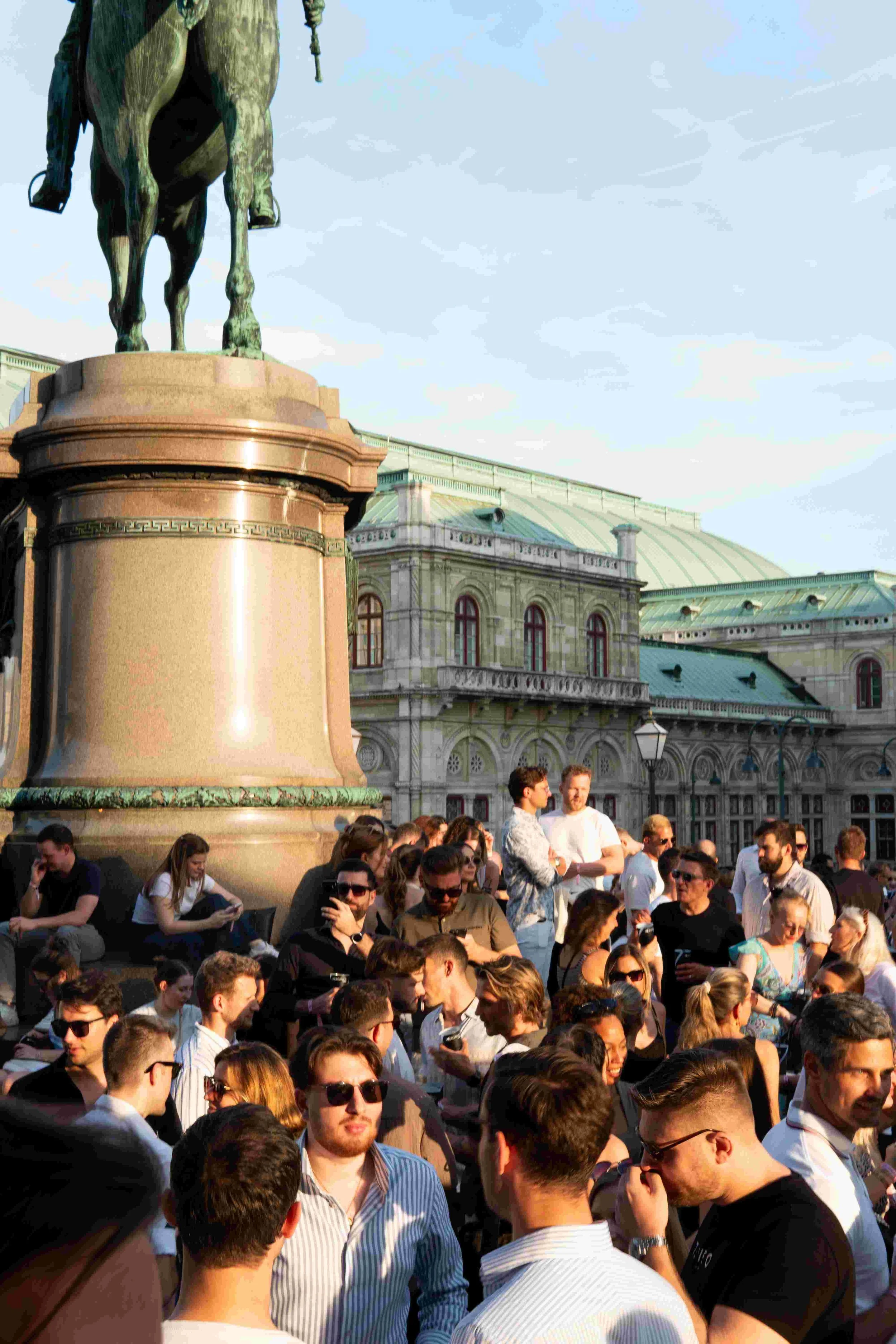 A large crowd of people gathered outdoors in front of a historic building with a green metal roof, and a prominent statue of a horse and rider on a pedestal. The sky is partly cloudy, and some people are interacting while others are using their phone