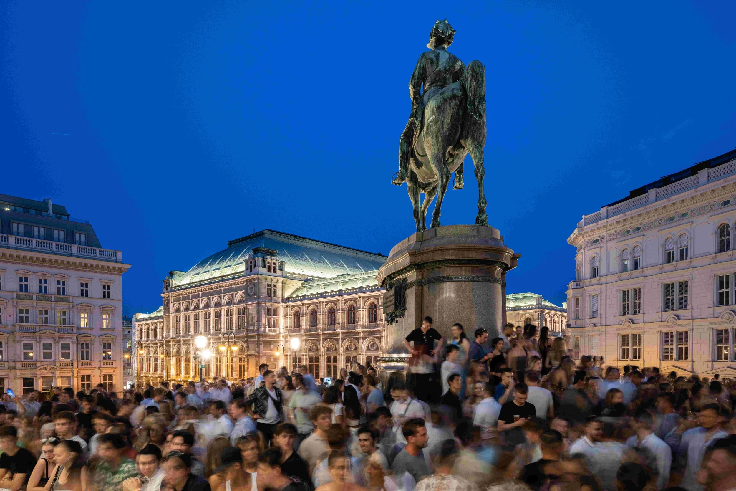 A busy city square at night with a crowd of people gathered and an elevated statue of a person on horseback against a blue sky. Buildings with ornate architecture are illuminated in the background.