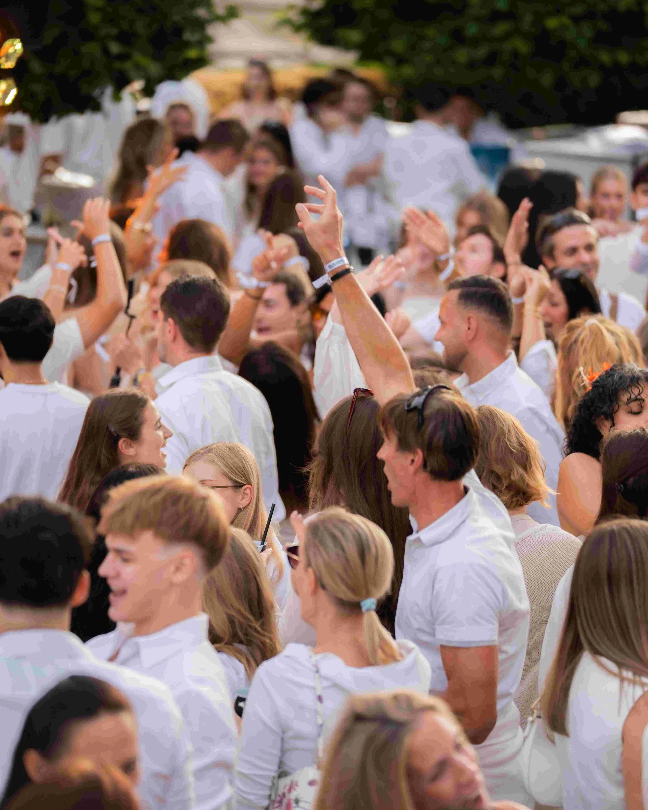 People gathered outdoors, dressed in white, participating in a celebration or event, with some raising their hands and engaging with each other.