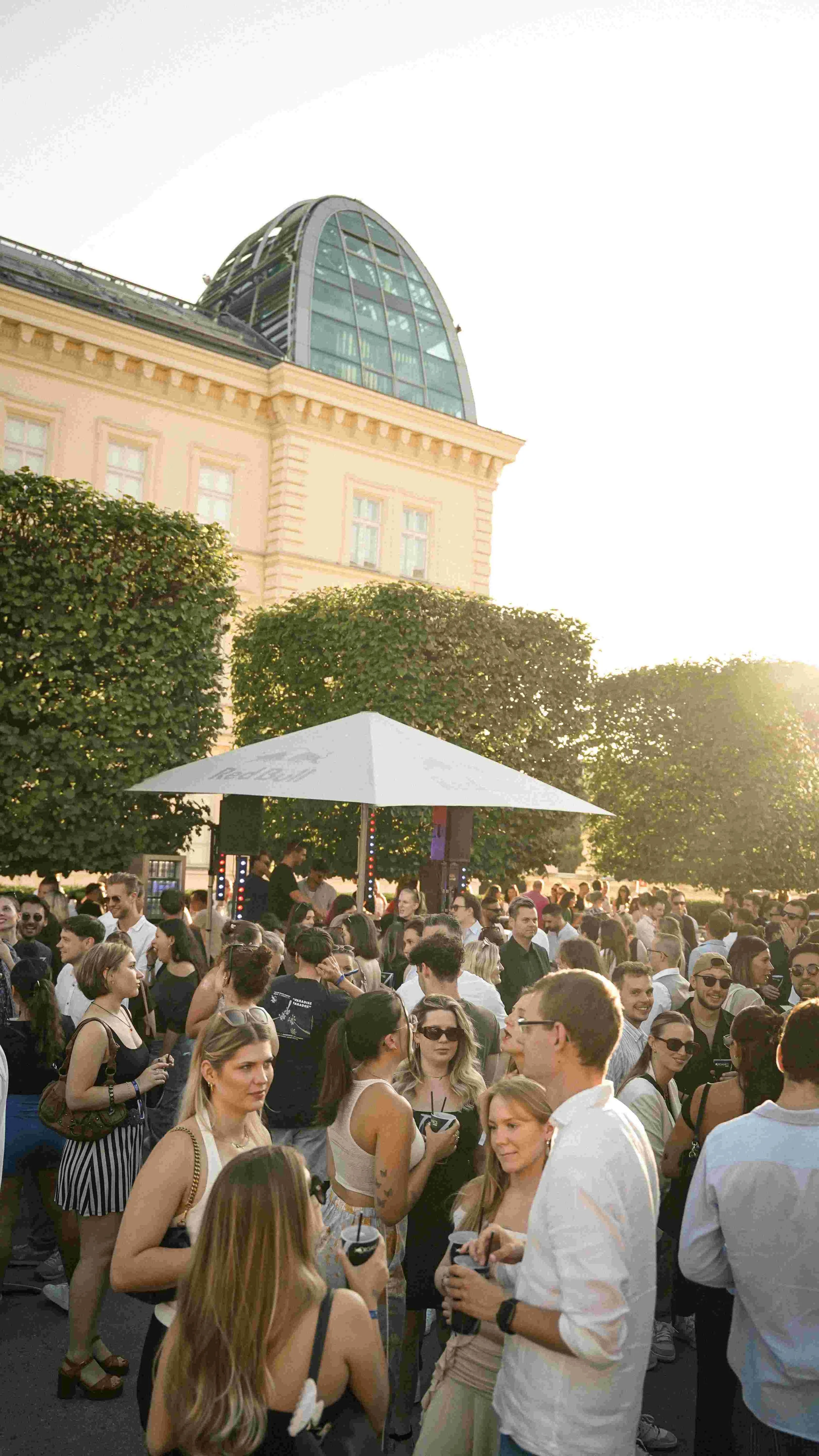 Crowd of people at an outdoor event with trees and a glass building in the background during sunset.