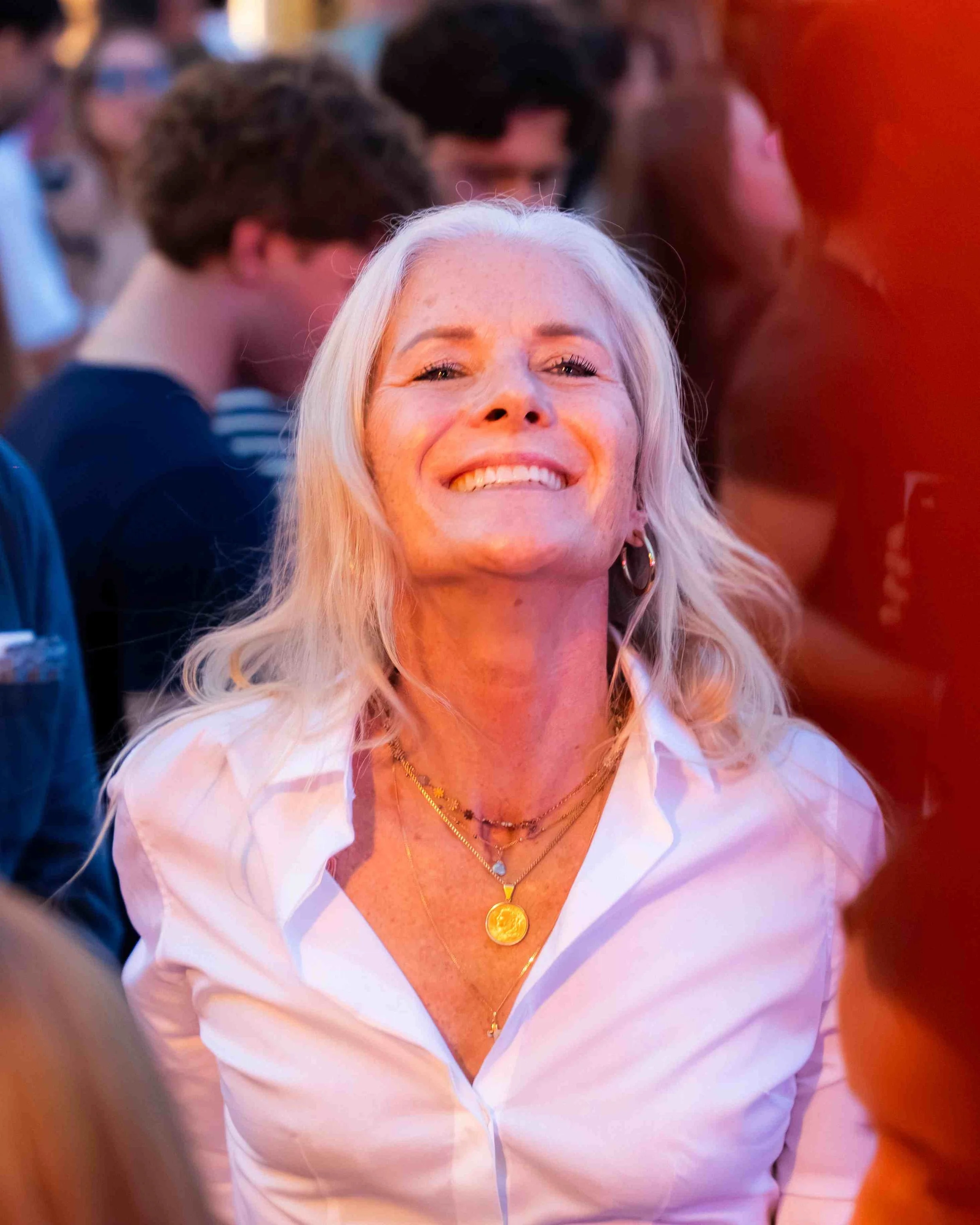 Smiling woman with long blonde hair wearing a white shirt and layered gold necklaces, at a lively social event with people in the background.