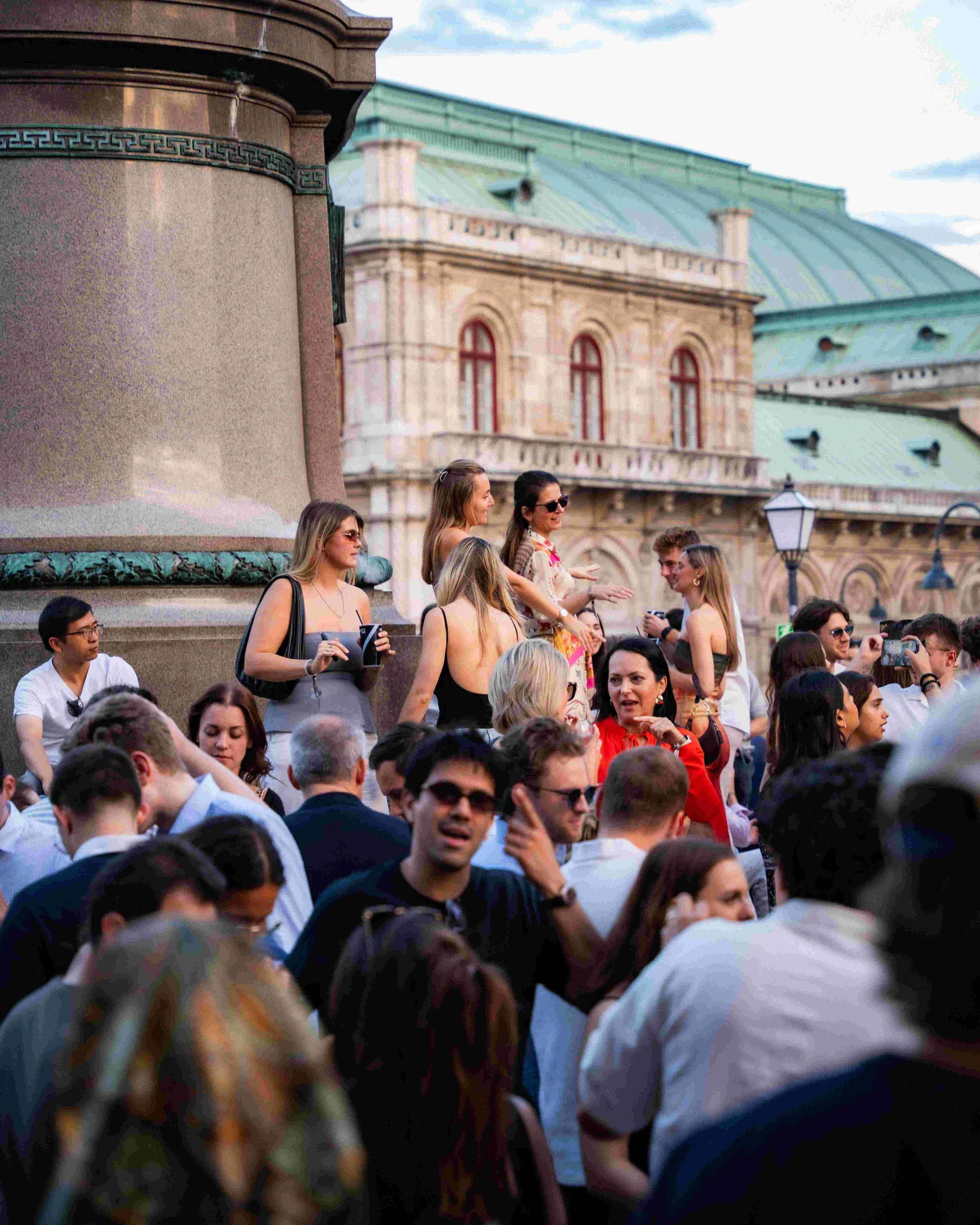 A crowd of people gathered outdoors in front of historic buildings with green rooftops and ornate windows, some standing and some sitting, during what appears to be a social event or gathering in an urban setting.