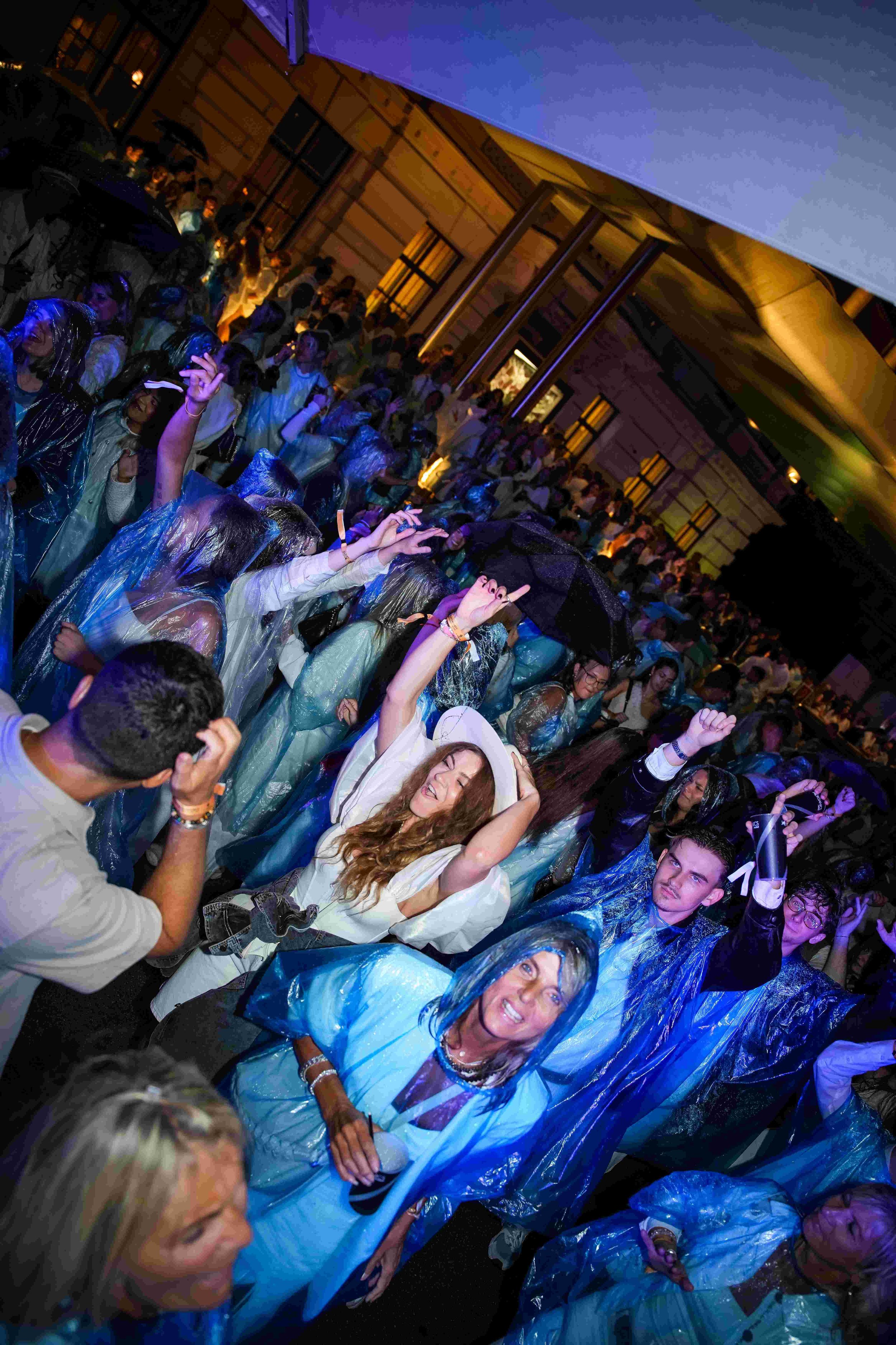 A large group of people attending an outdoor event at night, many wearing rain ponchos and holding umbrellas, with warm lighting and wooden structures in the background.