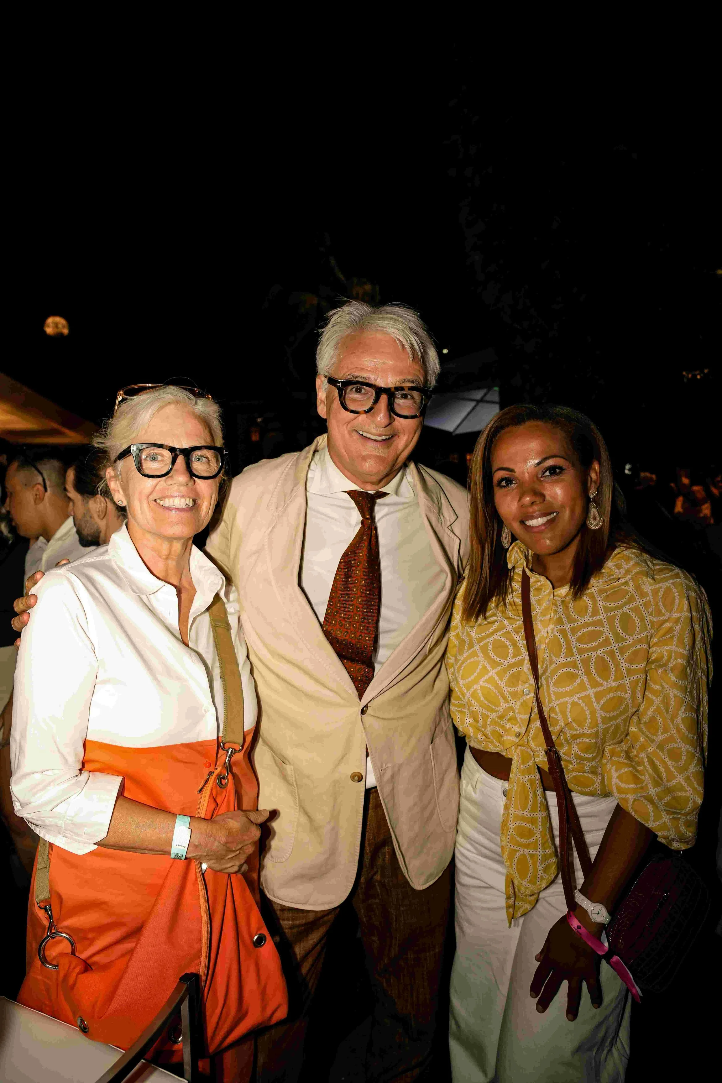 Four people, two women and two men, smiling at a nighttime outdoor event. They are dressed in semi-formal attire, with some wearing glasses and colorful accessories, standing close together.