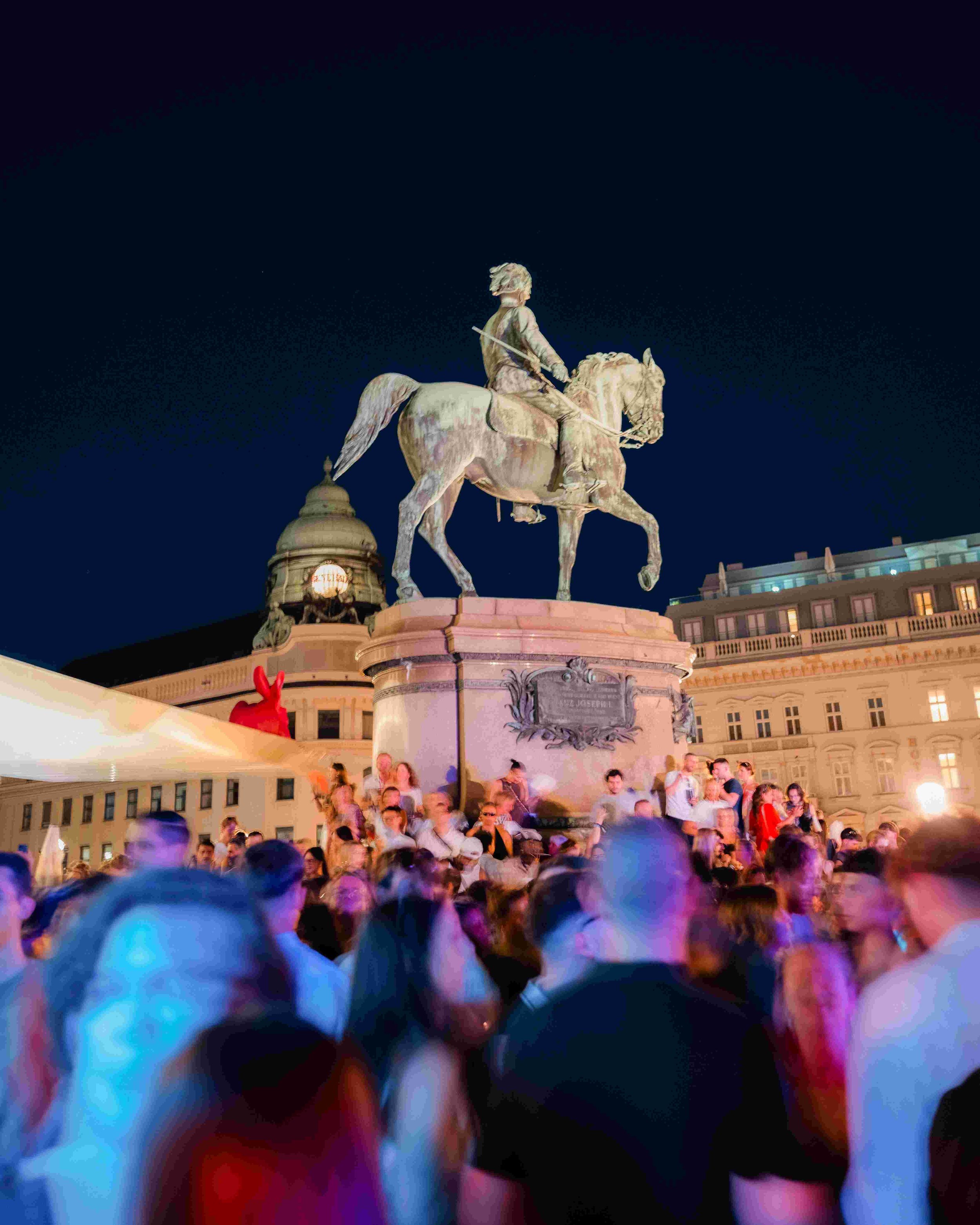 Nighttime scene of a large crowd gathered around a monument with a rider statue, with colorful lights and a historic building in the background.
