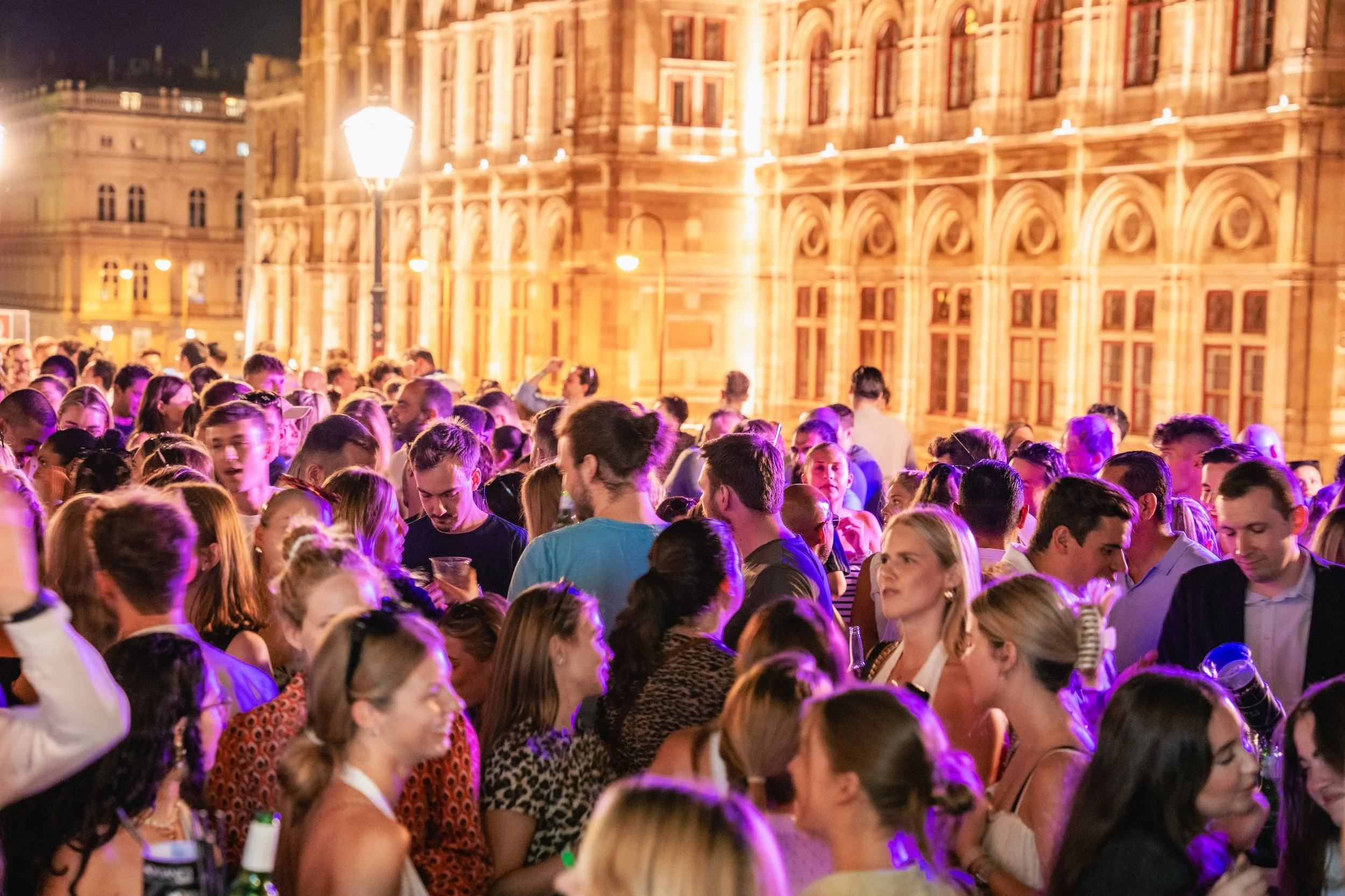 Crowd of people at an outdoor evening gathering in front of an illuminated historic building.