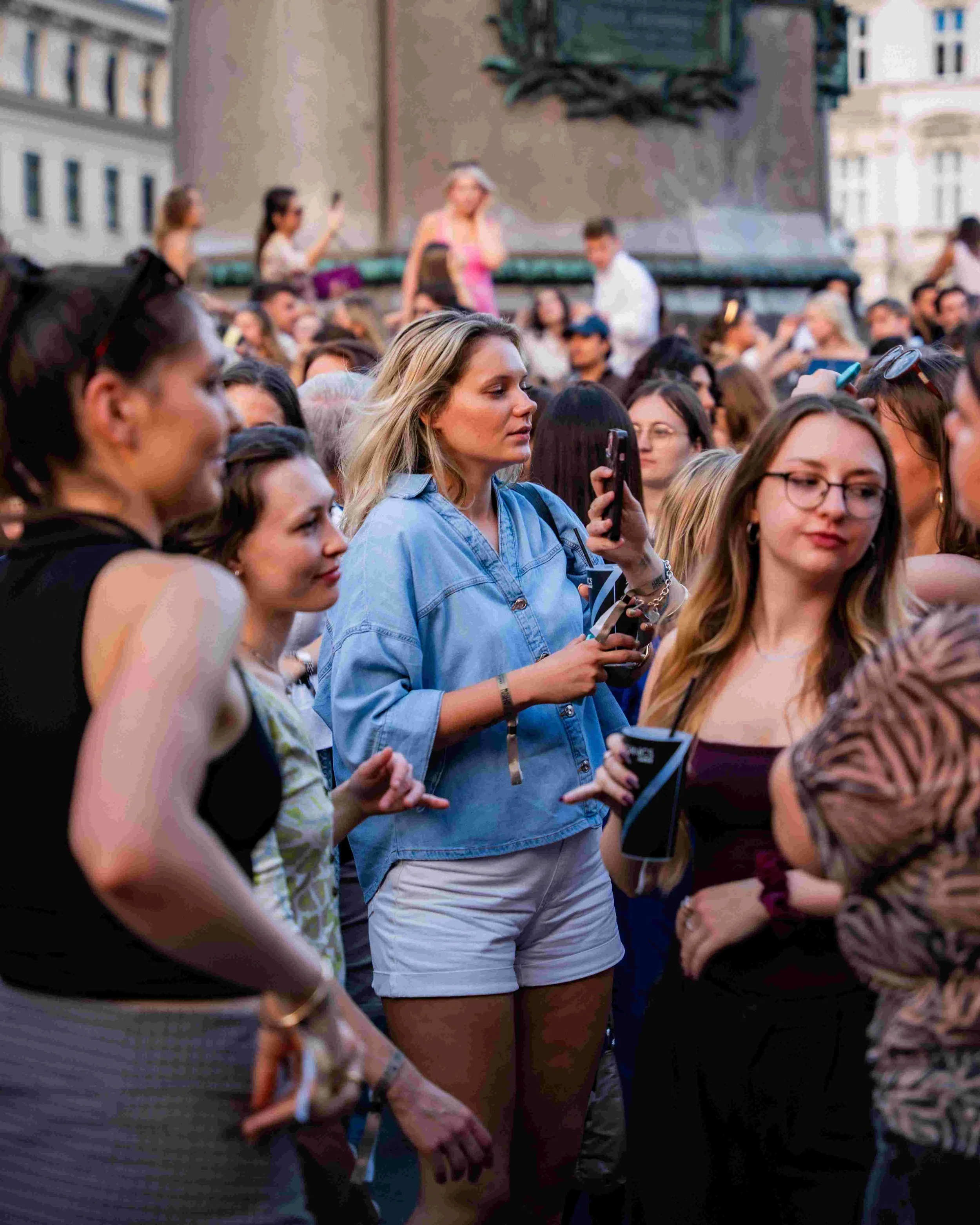 Crowd of people gathered in an outdoor urban setting, with some individuals taking photos or videos with their smartphones, and a woman in the center wearing a light blue denim shirt and white shorts, holding a phone.