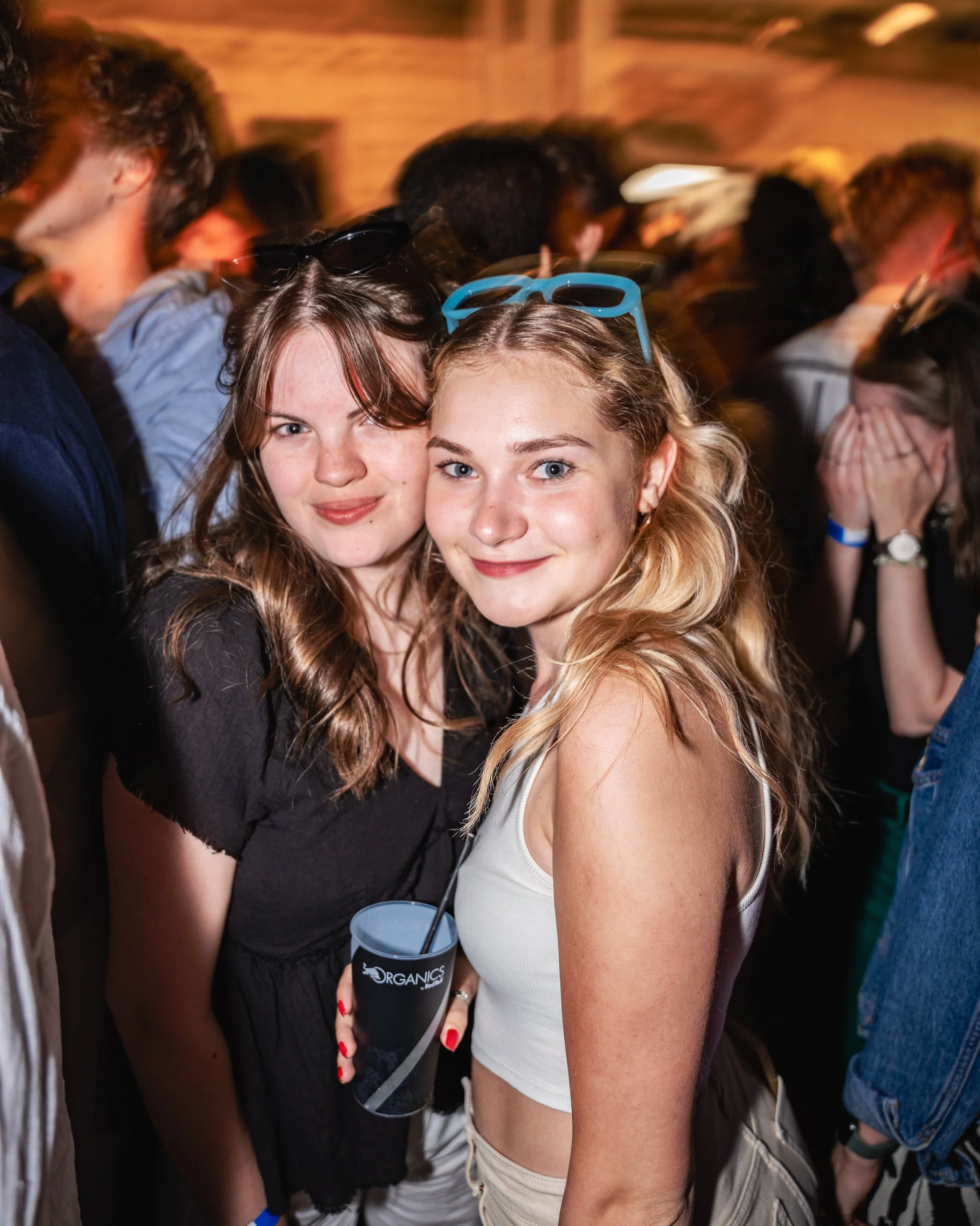 Two young women with long, wavy hair smiling for a selfie at a crowded event, with one holding a drink and sunglasses resting on her head.