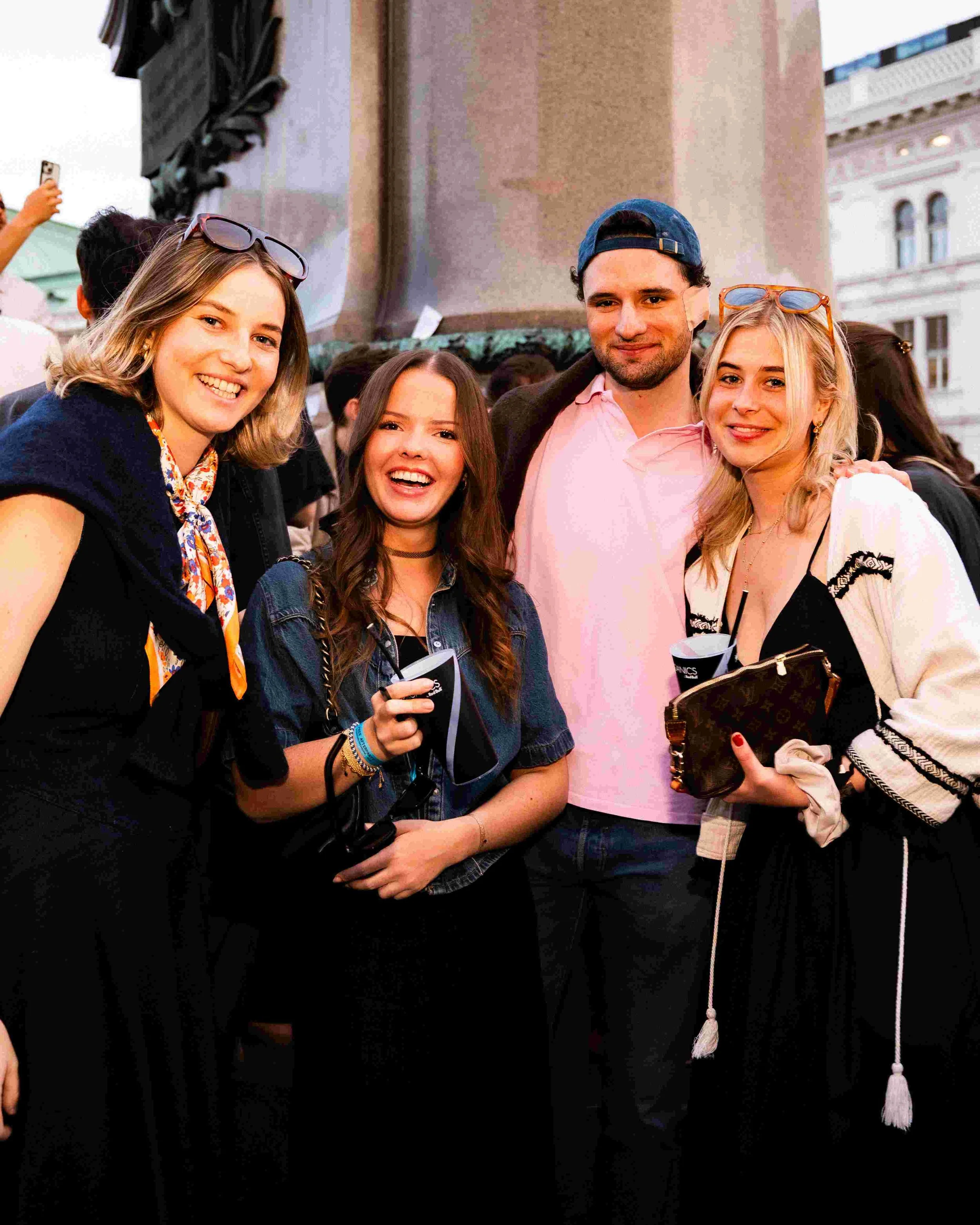 Group of four young adults taking a selfie at an outdoor event, with a large building and a crowd in the background.