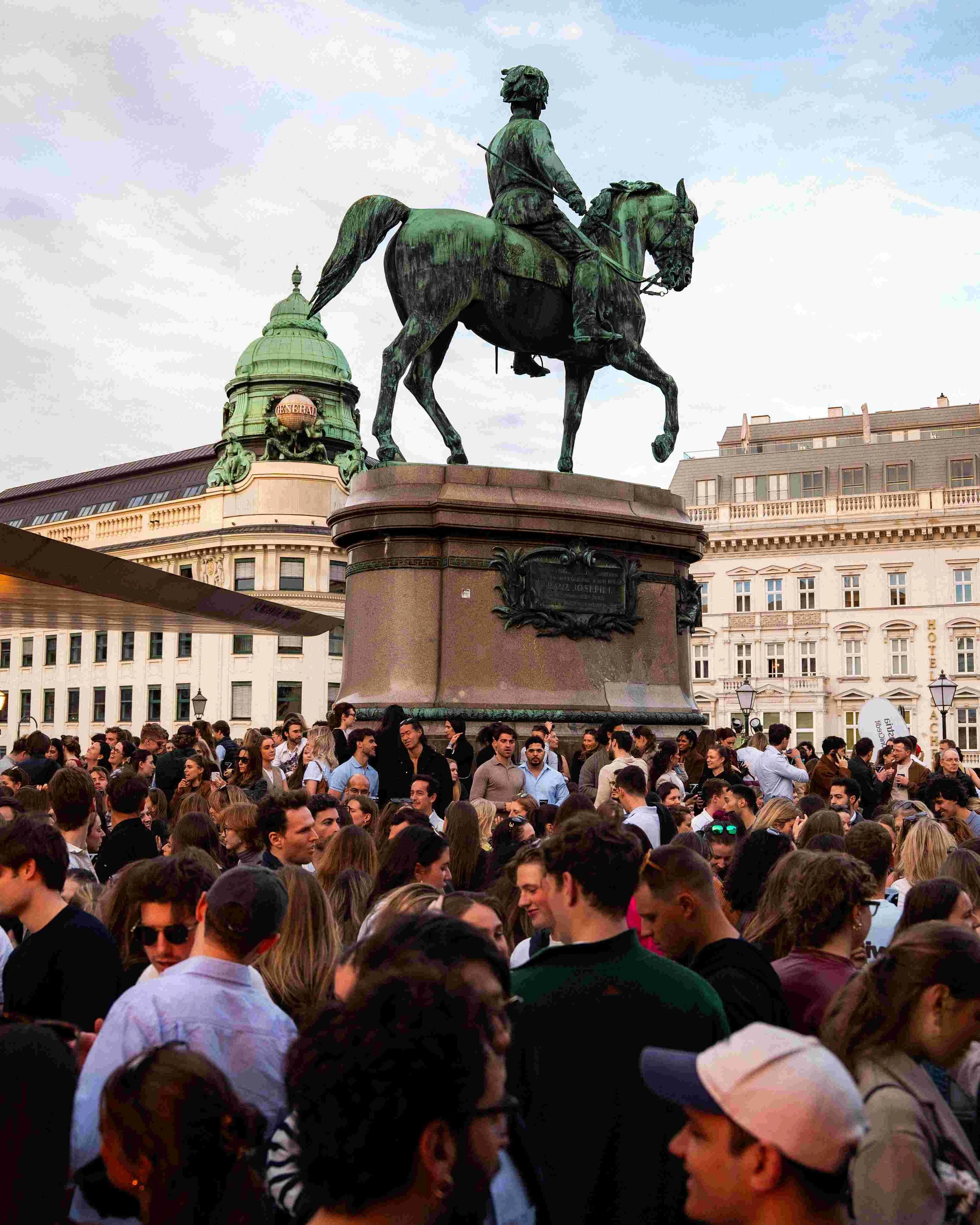 Crowd of people gathered around an equestrian statue of a man in a city square, with historic buildings in the background.