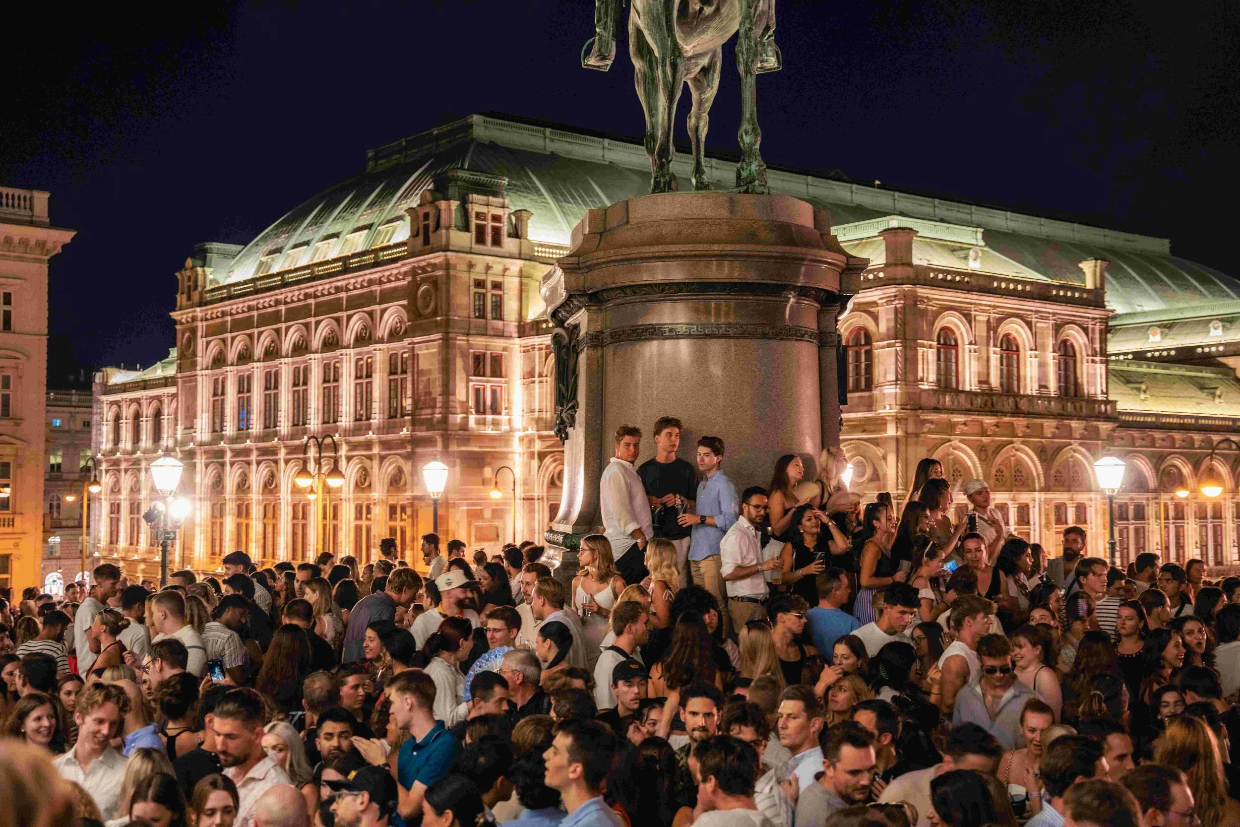A large crowd of people gathered in an outdoor square at night, with a historic building featuring ornate architecture and illuminated windows in the background. A statue on a pedestal is visible in the foreground.
