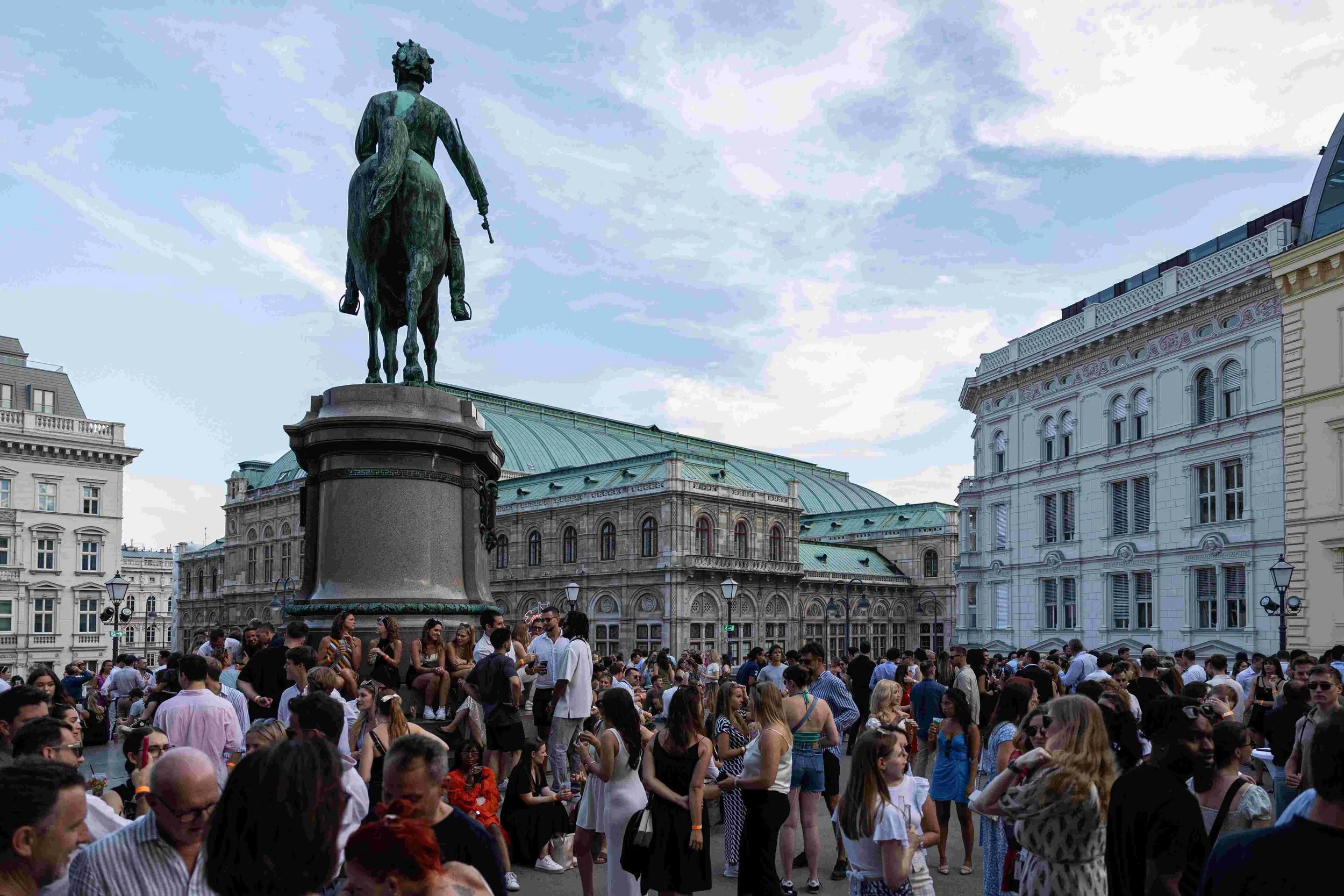 Crowd of people gathering in a city square near a statue of a man on a horse, with historic buildings and cloudy sky in the background.