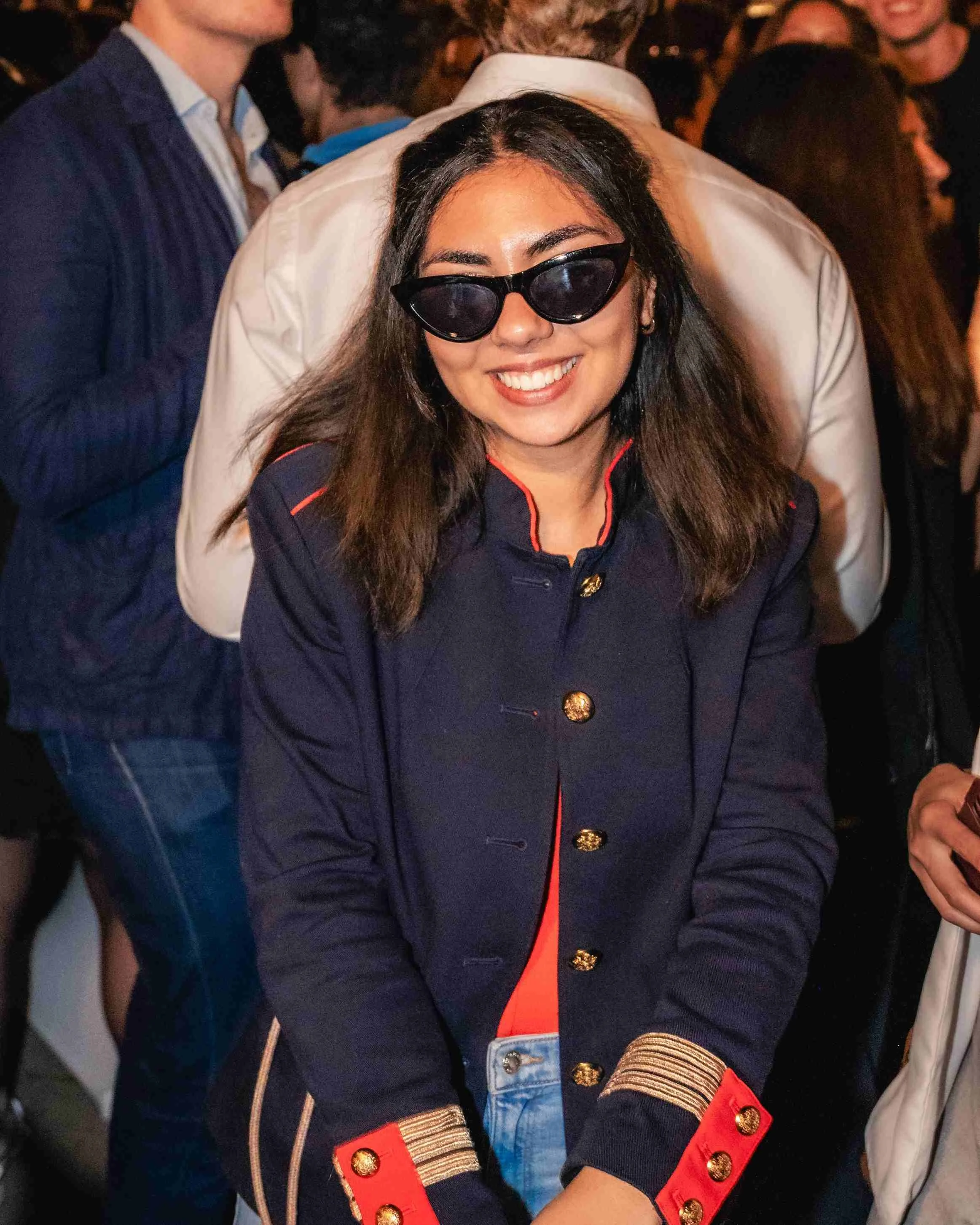 A smiling woman wearing black sunglasses and a navy blue jacket with red accents and gold buttons, at a crowded event.