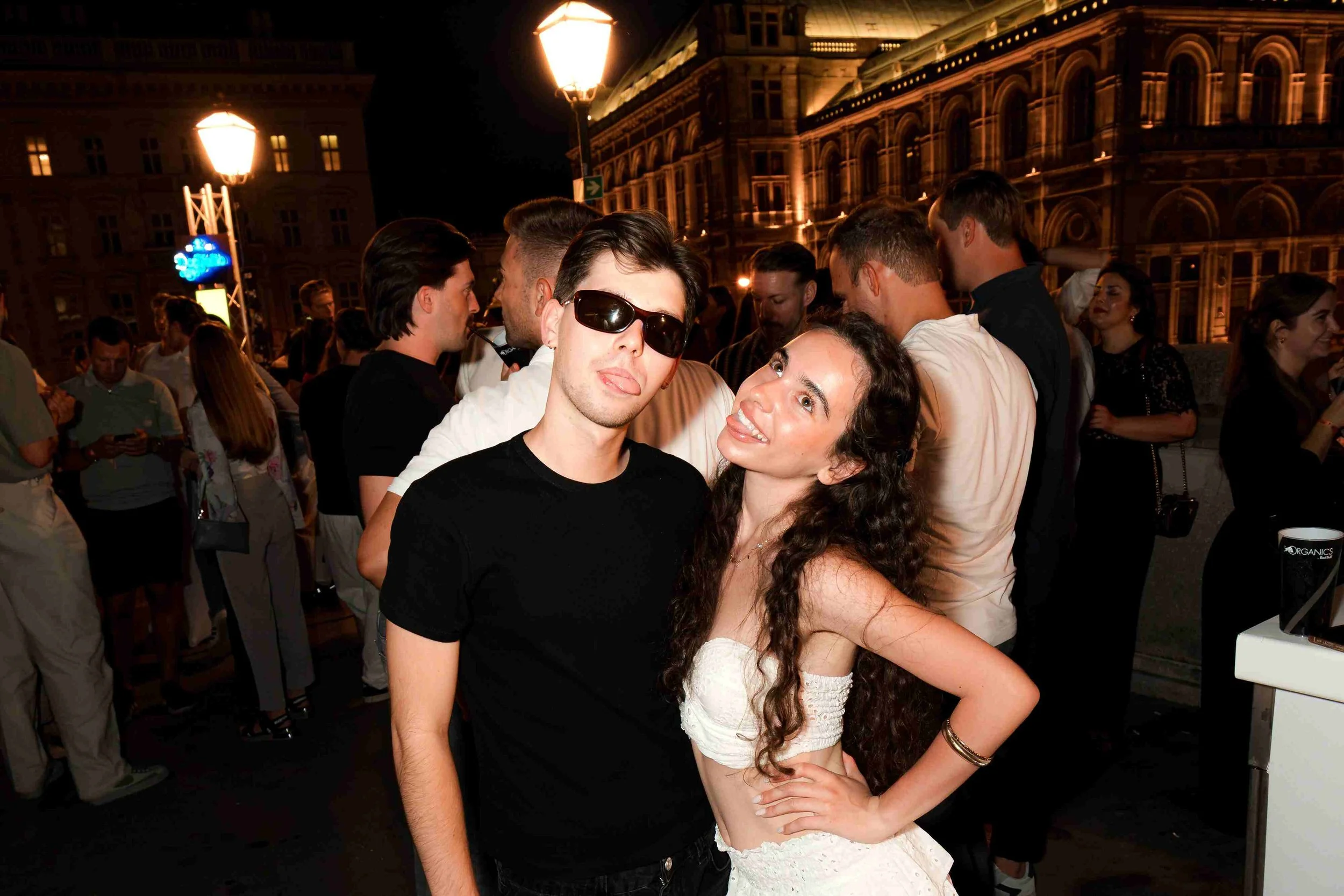A young man wearing sunglasses and a young woman with long curly hair in a white dress pose playfully together at an outdoor night event, with a crowd of people and illuminated historic buildings in the background.