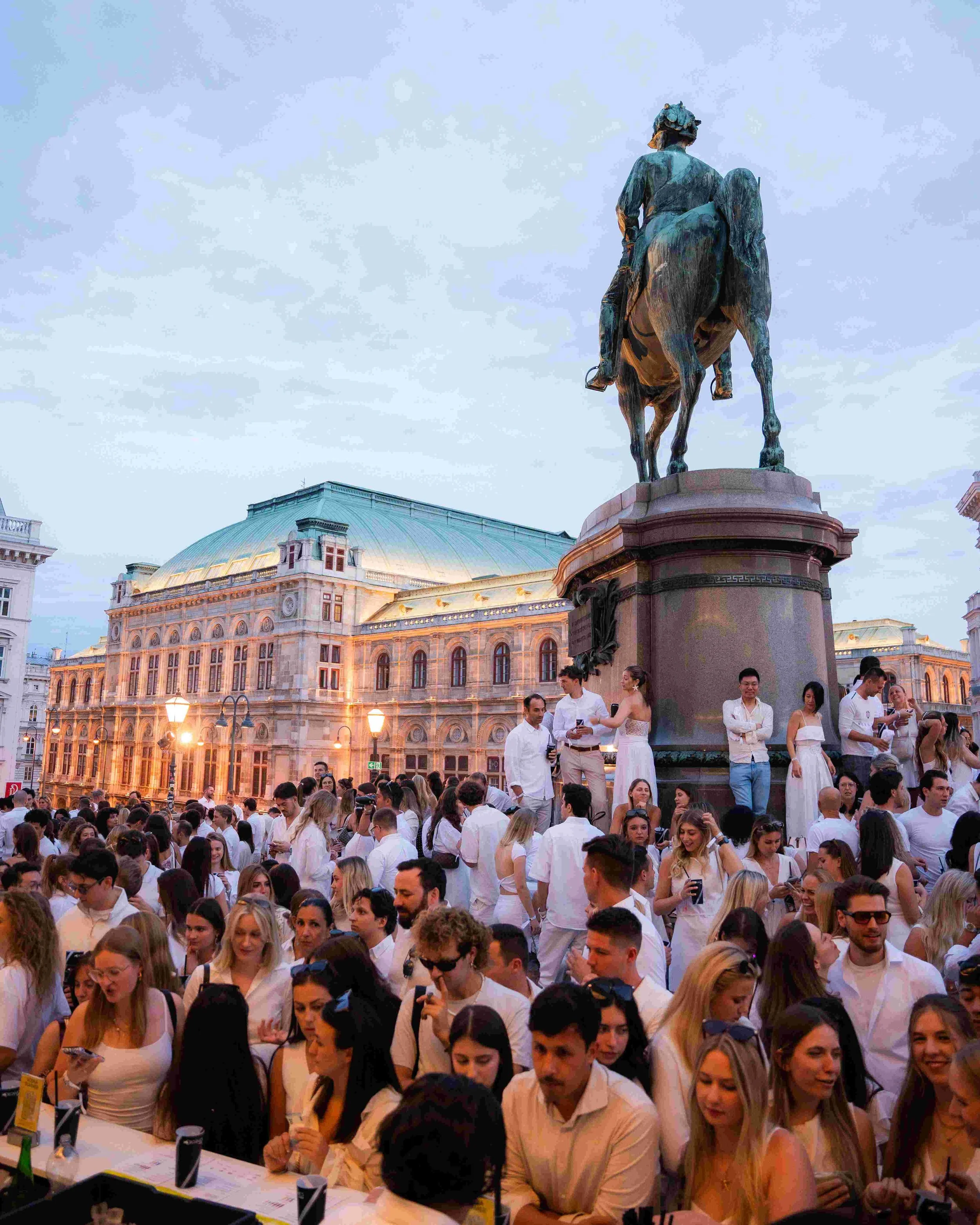 Large crowd of people gathered outdoors in front of historic building with a domed roof, around a table with numbered bottles and papers, during evening or early night.