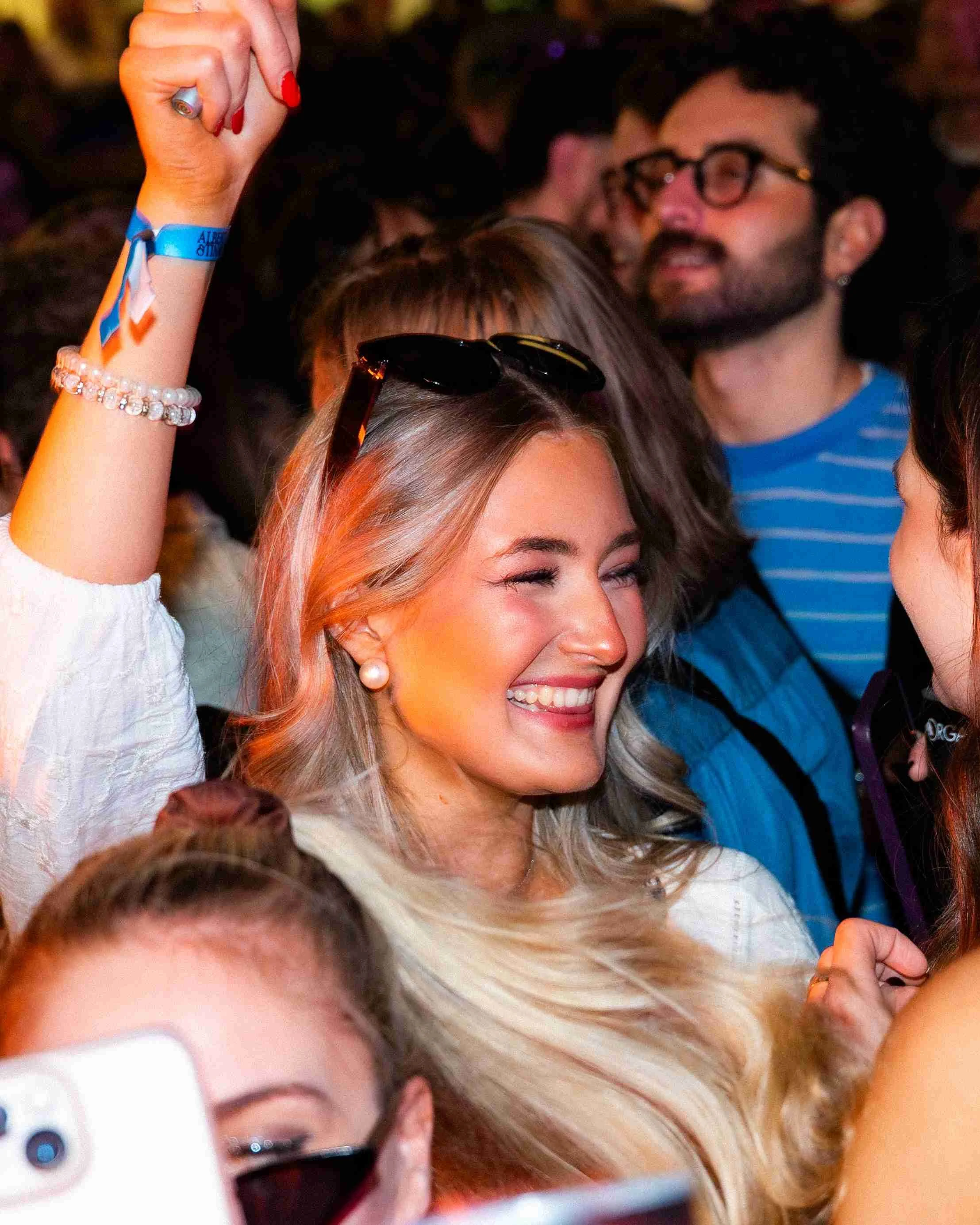 A woman with blonde hair and pearl earrings smiling, wearing sunglasses on her head, in a crowd at an event.
