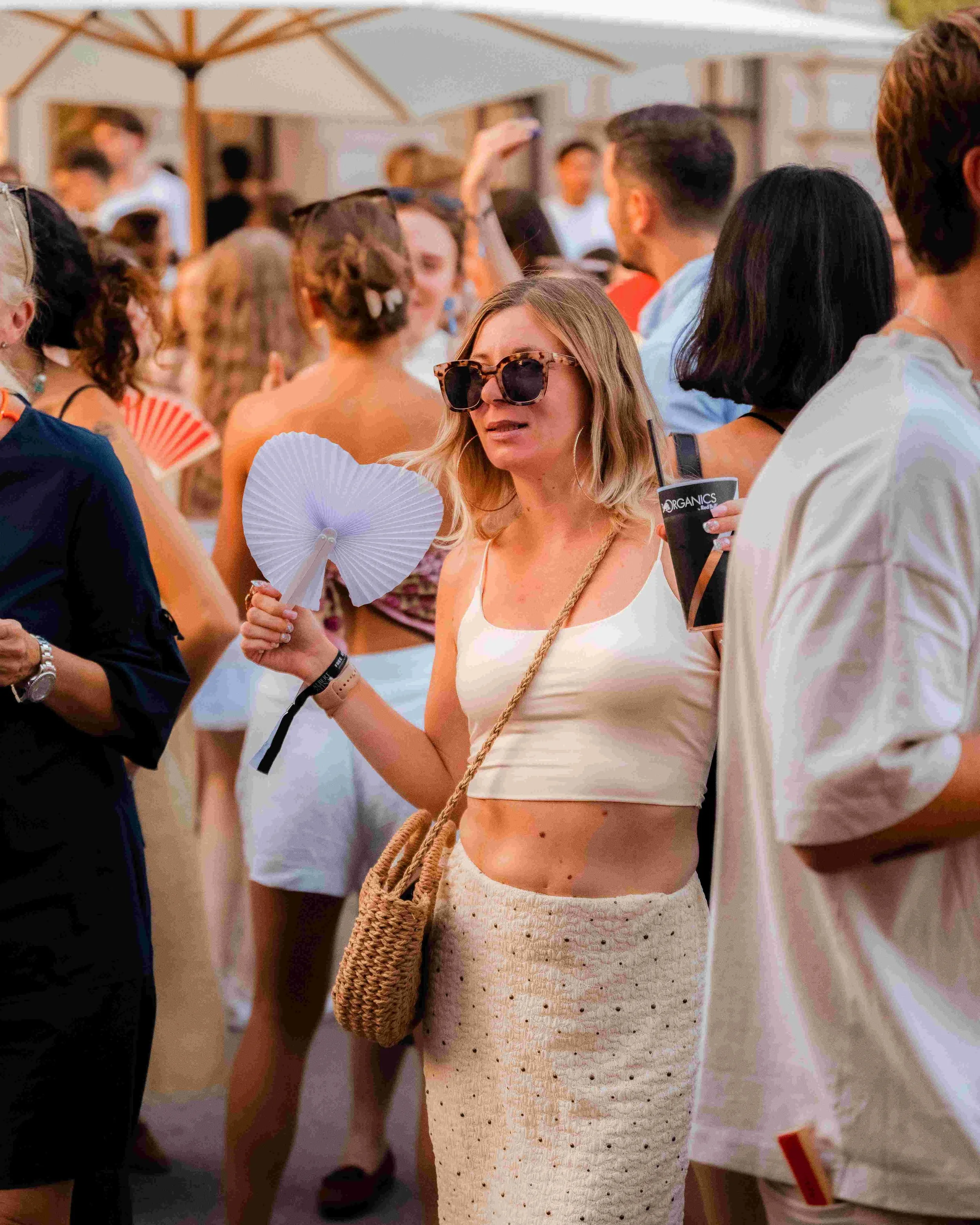 A group of people at an outdoor social gathering under a large white patio umbrella, with a woman in the foreground wearing sunglasses, a white tank top, and a black wristband, holding a paper fan and a drink, engaging in conversation.