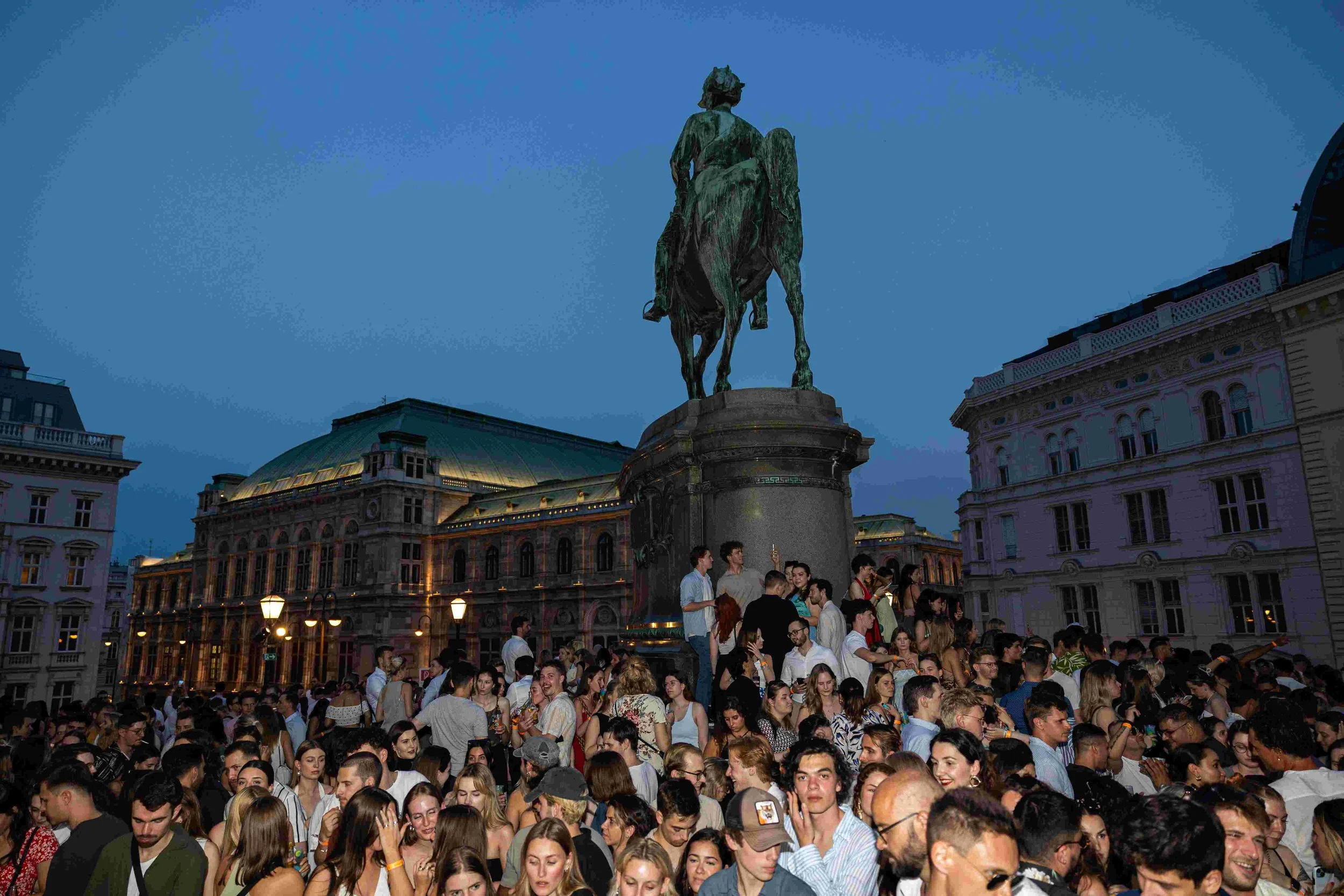 Crowd of people gathered at an outdoor event during evening with historic buildings and a large equestrian statue in the background.