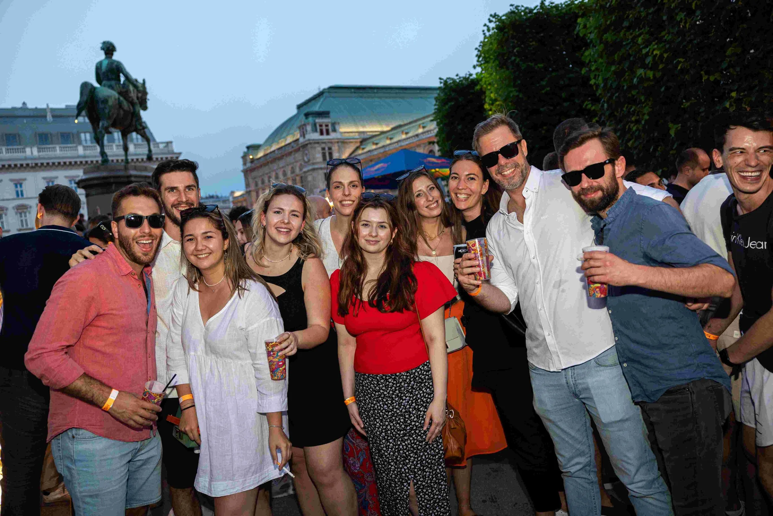 A group of young adults smiling and posing for a photo at an outdoor event, with historic buildings and a statue in the background.