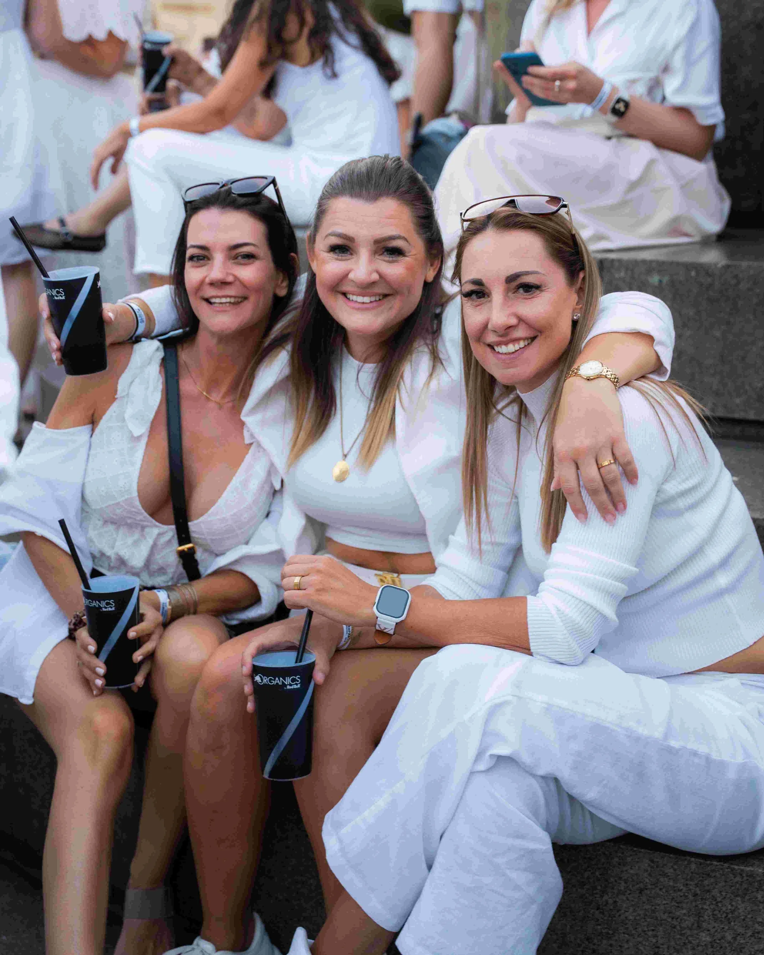 Three smiling women sitting outdoors, hugging each other, holding black cups with straws, and wearing white clothing. Two women have sunglasses on their heads, and they are enjoying a casual gathering.