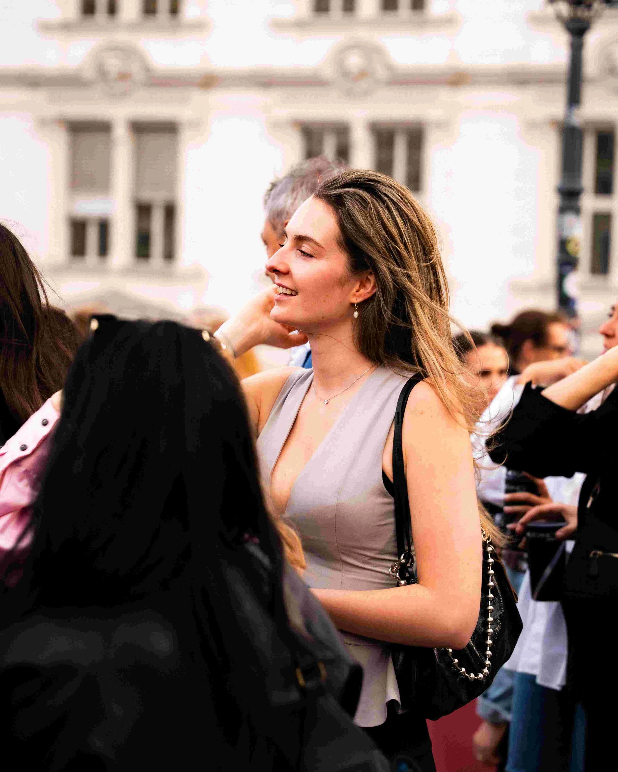 A young woman with long brown hair, smiling with eyes closed, wearing a light gray sleeveless top, a pearl earring, and a beaded purse, in a crowd of people outdoors.