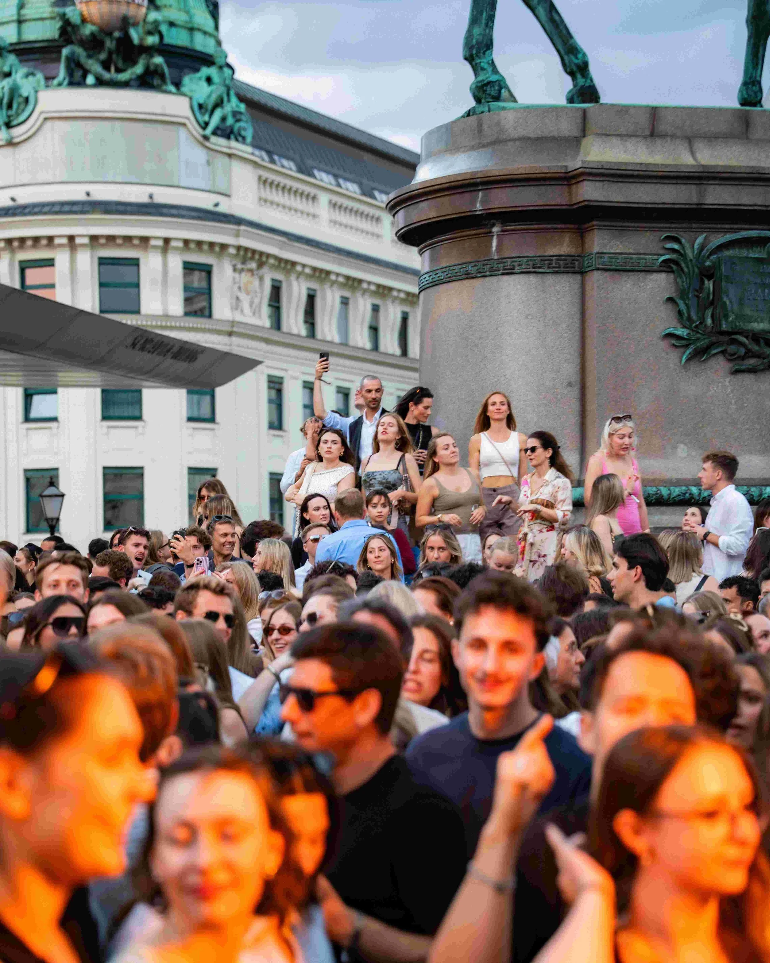 Crowd of people gathered outdoors with historic buildings in the background, some taking photos, under a cloudy sky.