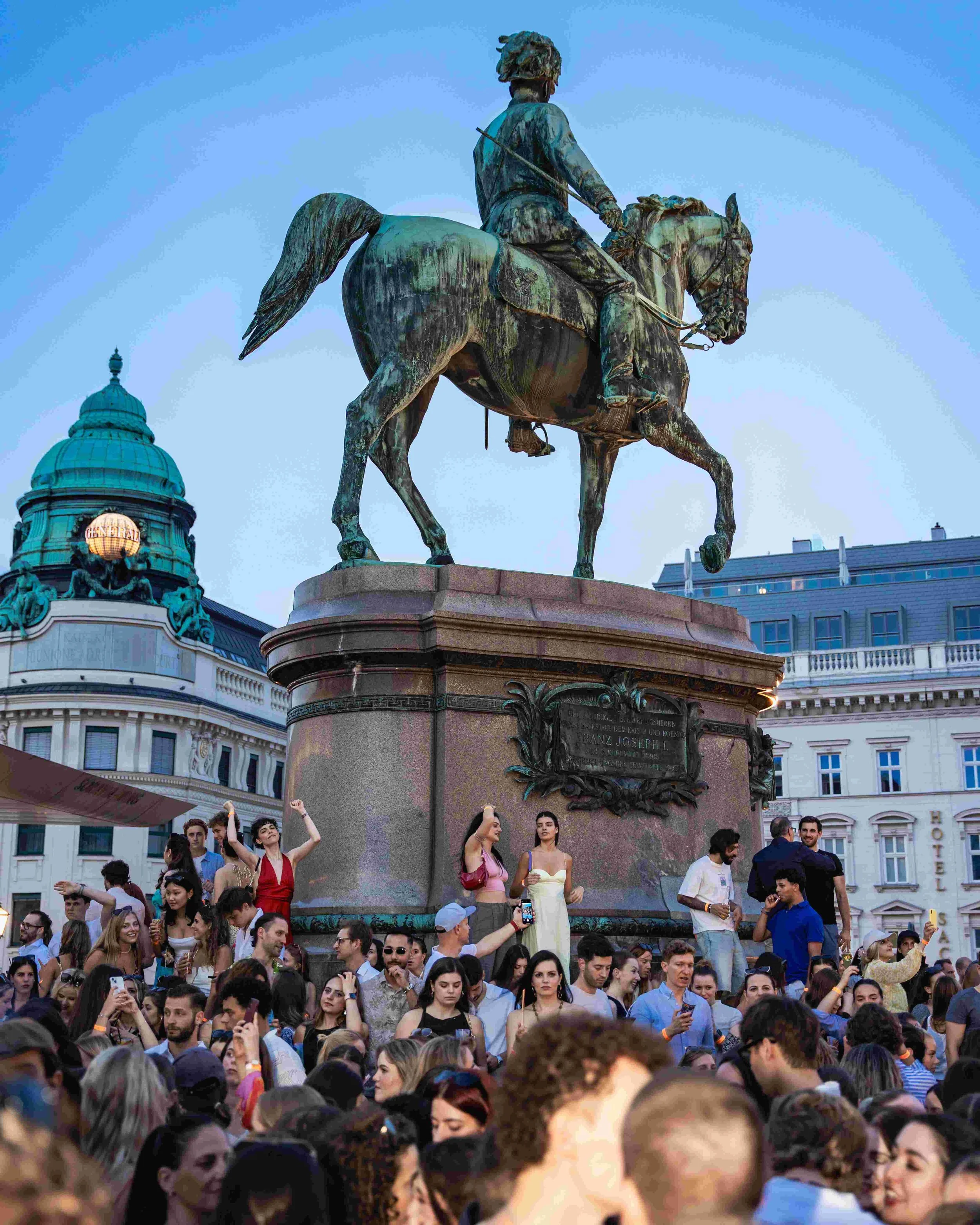 Crowd of people gathered around a statue of a man on horseback in an urban square, with historic buildings and a domed tower in the background.