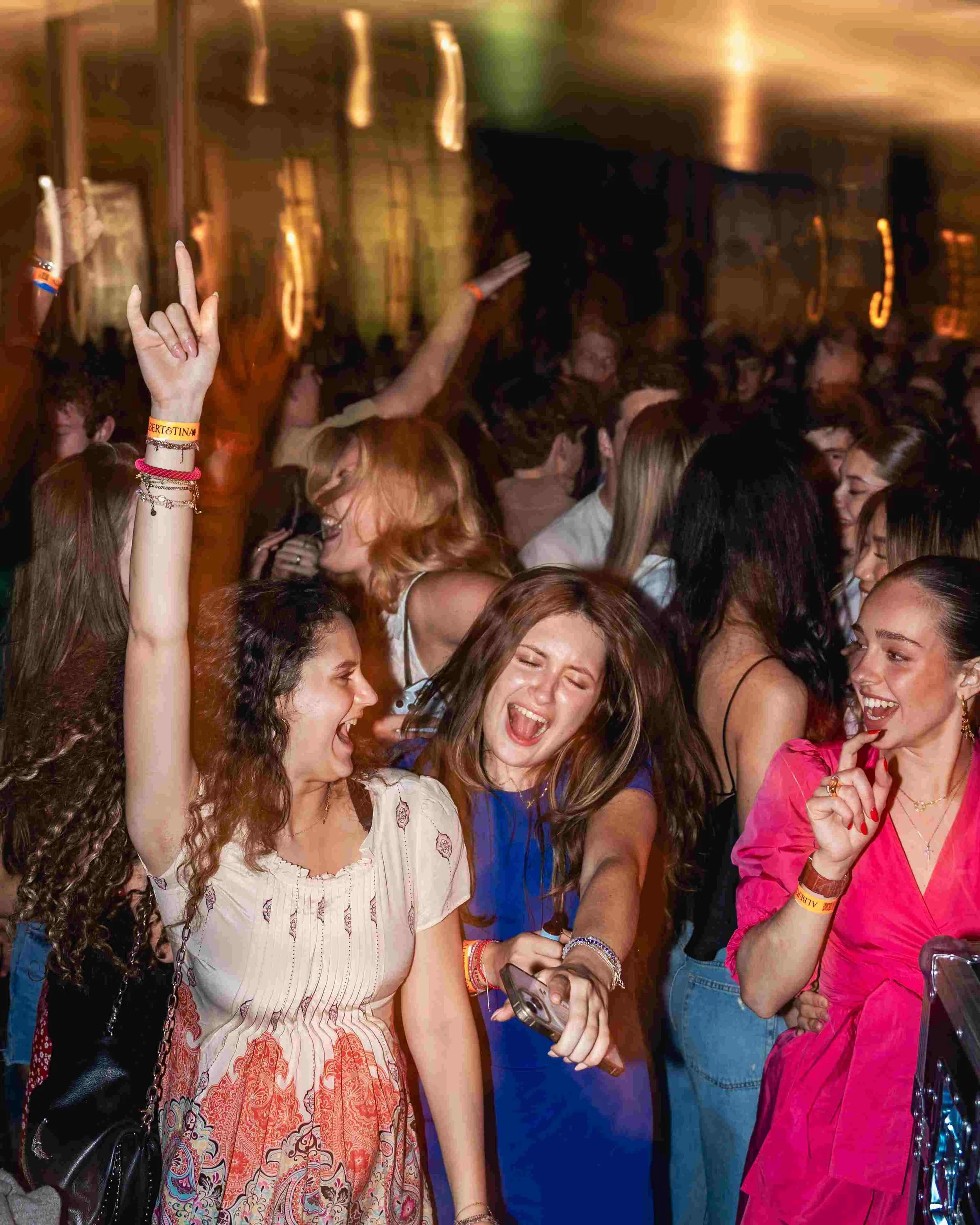Crowd of people at a party or concert, some raising their hands, smiling, wearing colorful outfits, and accessories.