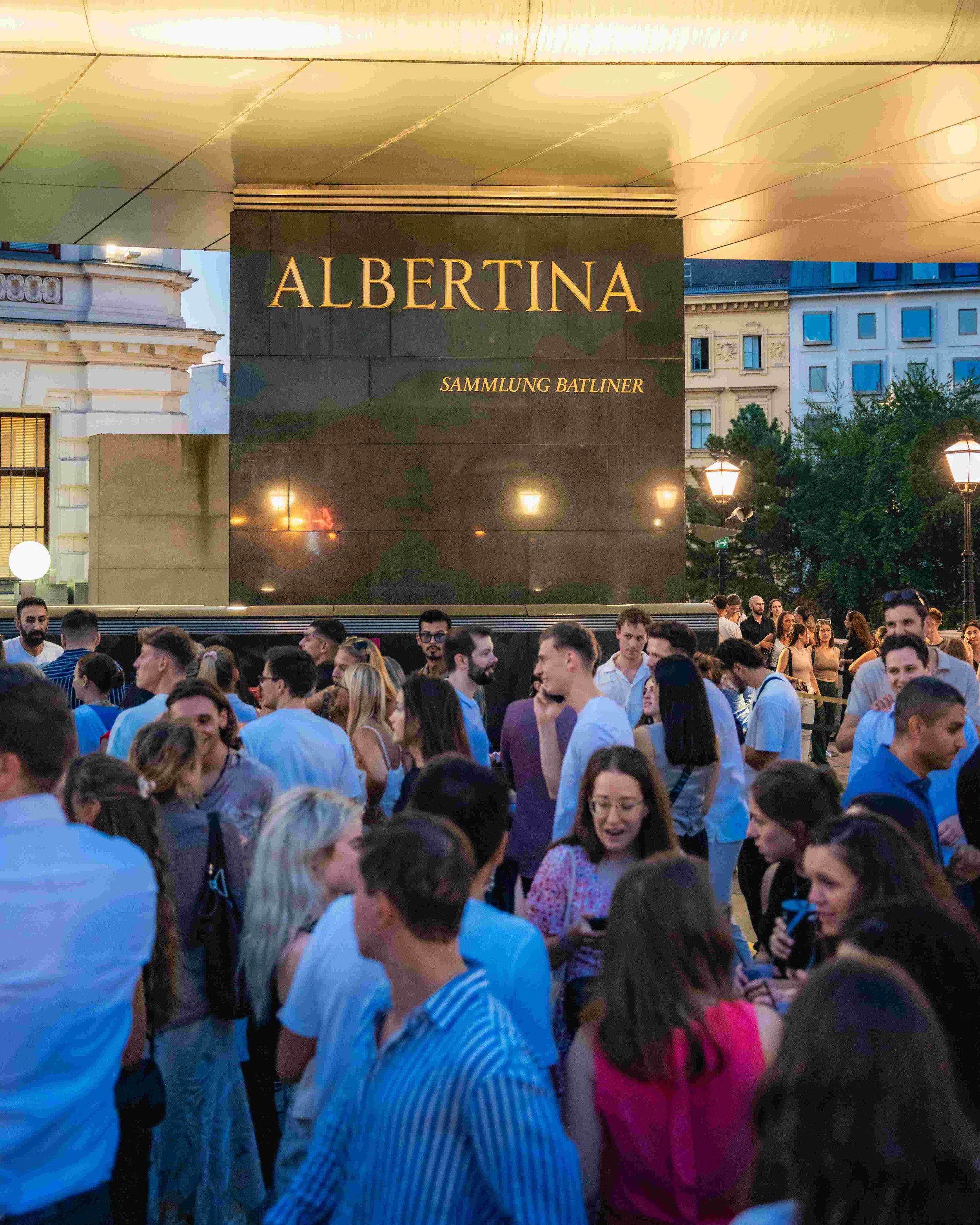 A large crowd of people gathered outdoors near a building with a modern, overhanging roof, and a sign that reads 'ALBERTINA SAMMLUNG BATLINER.' The crowd consists of men and women of various ages, some looking at their phones, and some engaged in con