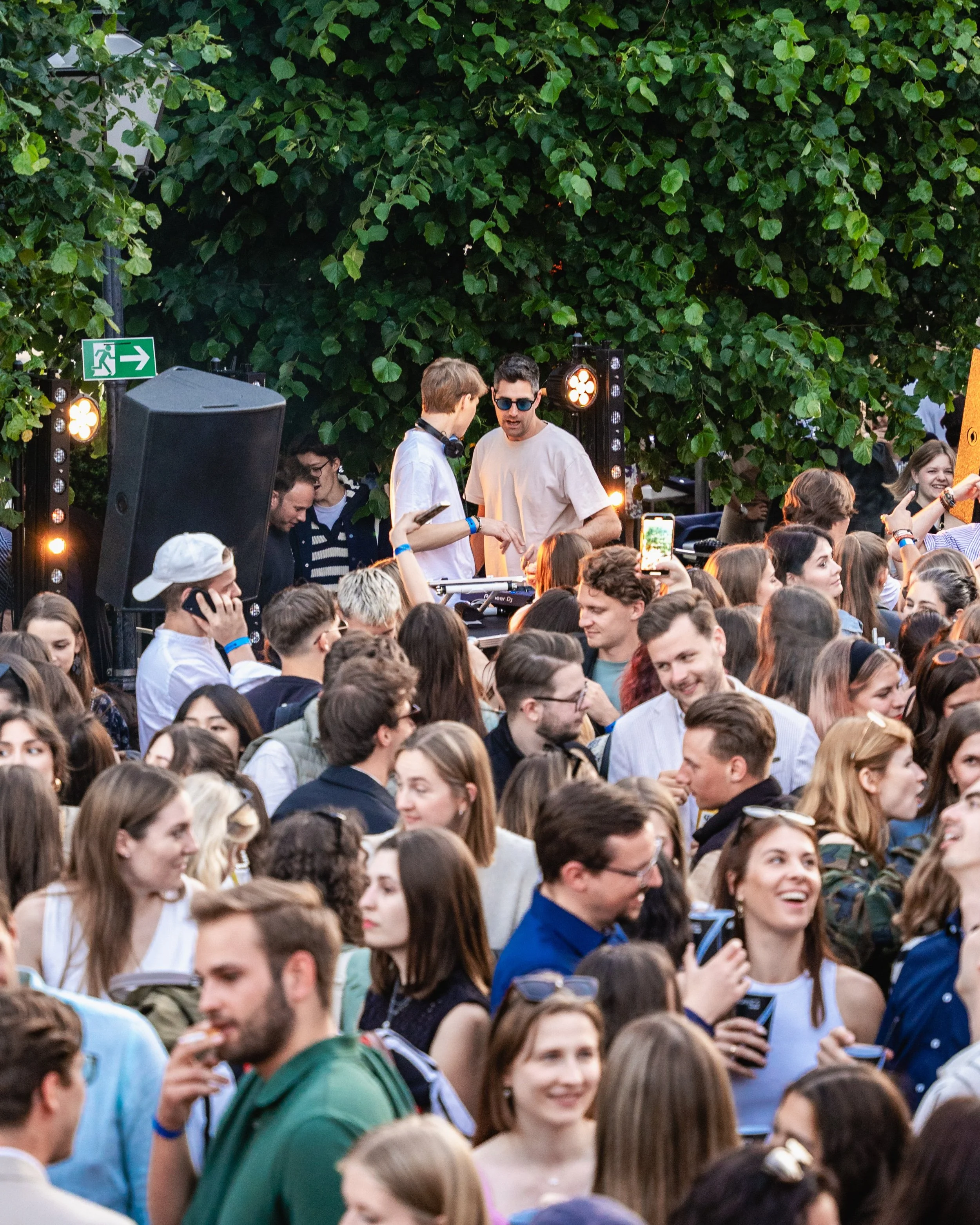 Crowd of people at an outdoor event with a DJ playing music. Some people are taking photos and videos, and there are bright stage lights and green foliage in the background.