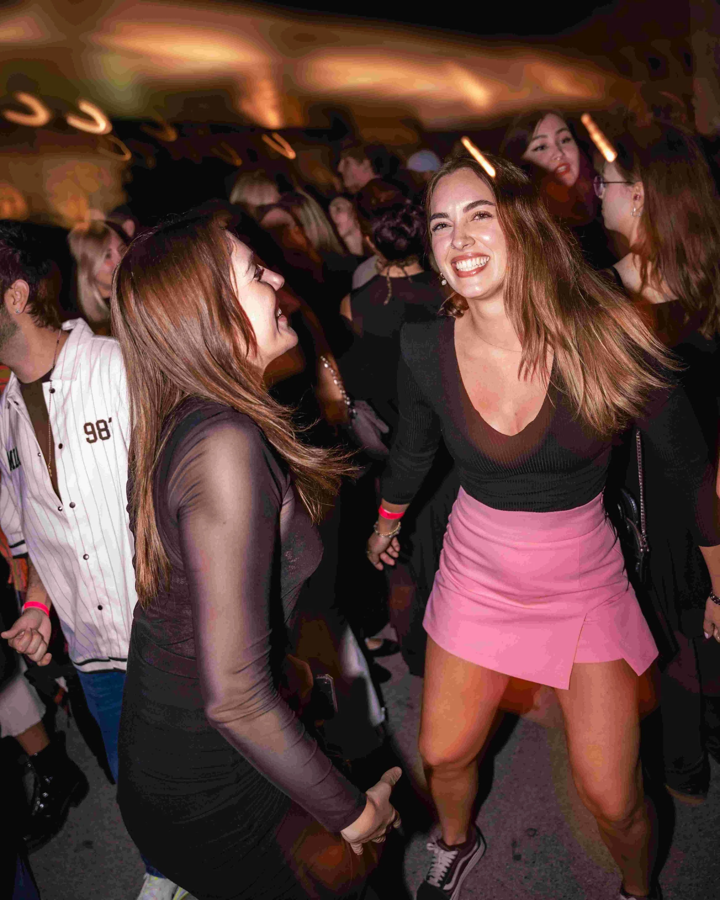 A woman in a black top and pink skirt dancing and smiling at a crowded party or club, surrounded by other people.