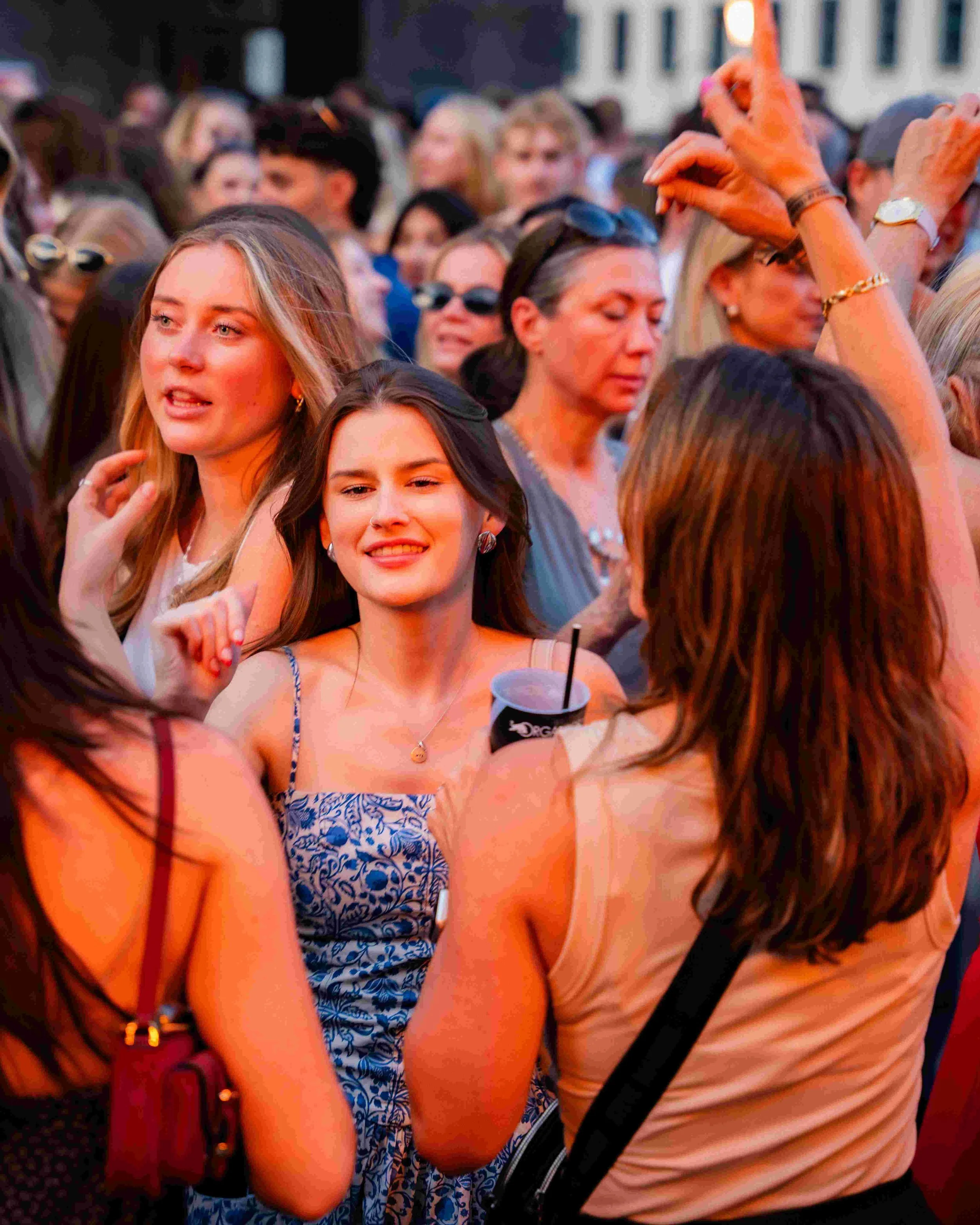 A crowd of people at an outdoor event, with some dancing and socializing. Two women in the foreground are smiling and enjoying themselves. One woman is holding a beverage in a cup.