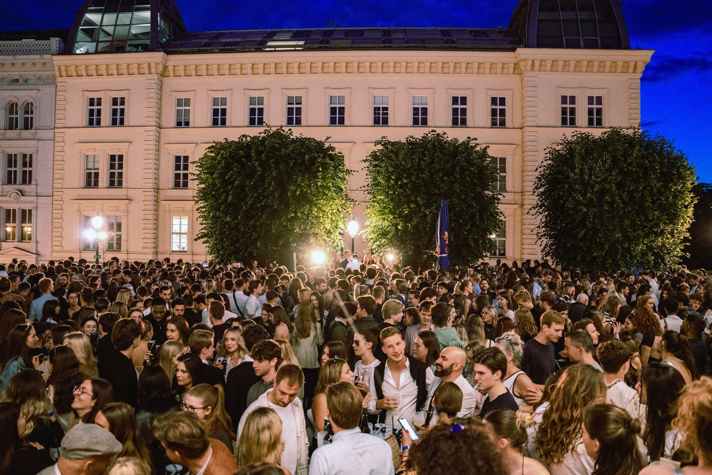 Large crowd of people gathered outdoors at night in front of a historic building with trees and streetlights.