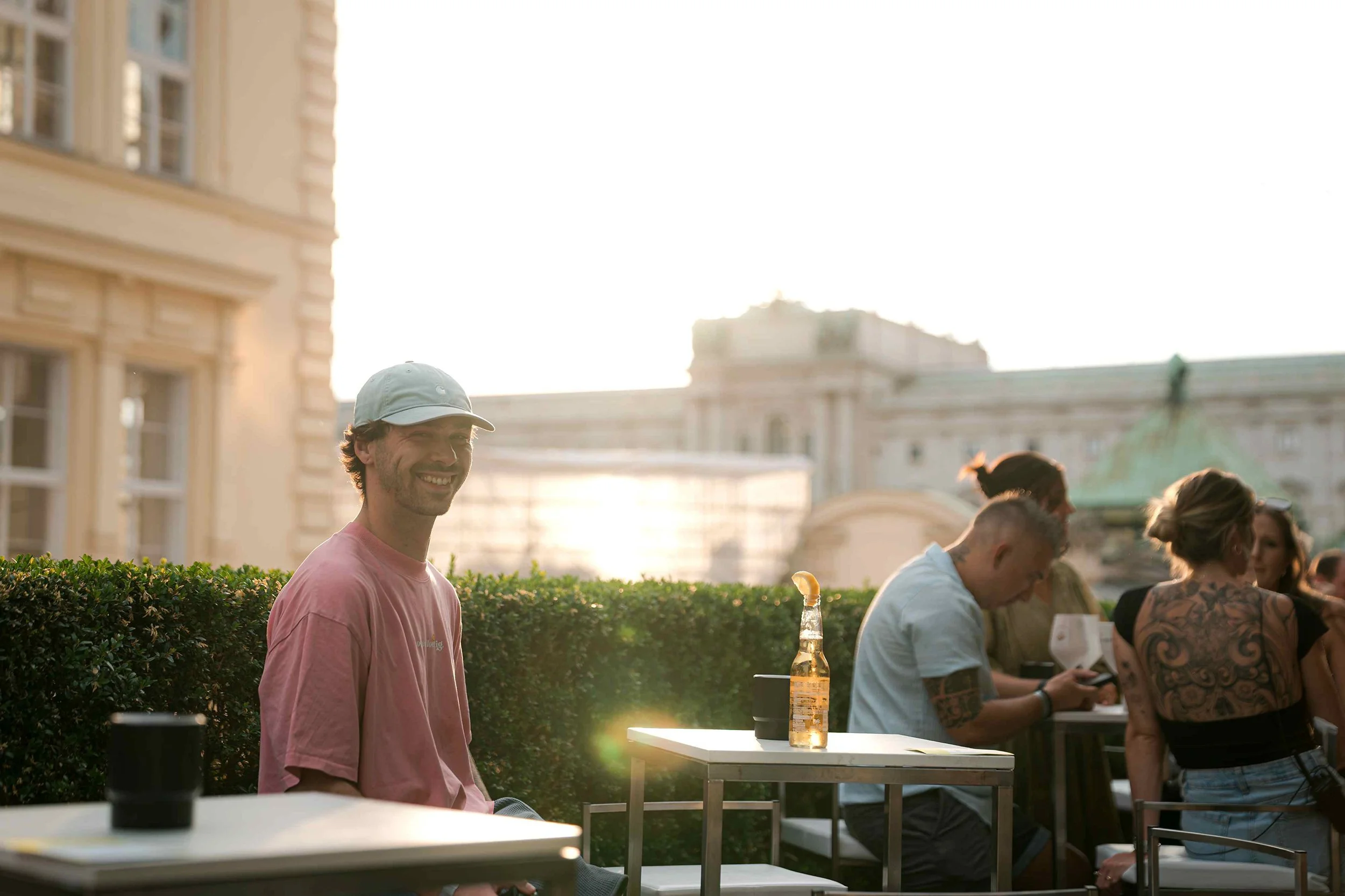 A man in a pink shirt and white cap smiling while sitting outdoors in a cityscape with buildings in the background, with a group of people gathered at tables nearby, and the sunlight creating a warm glow.