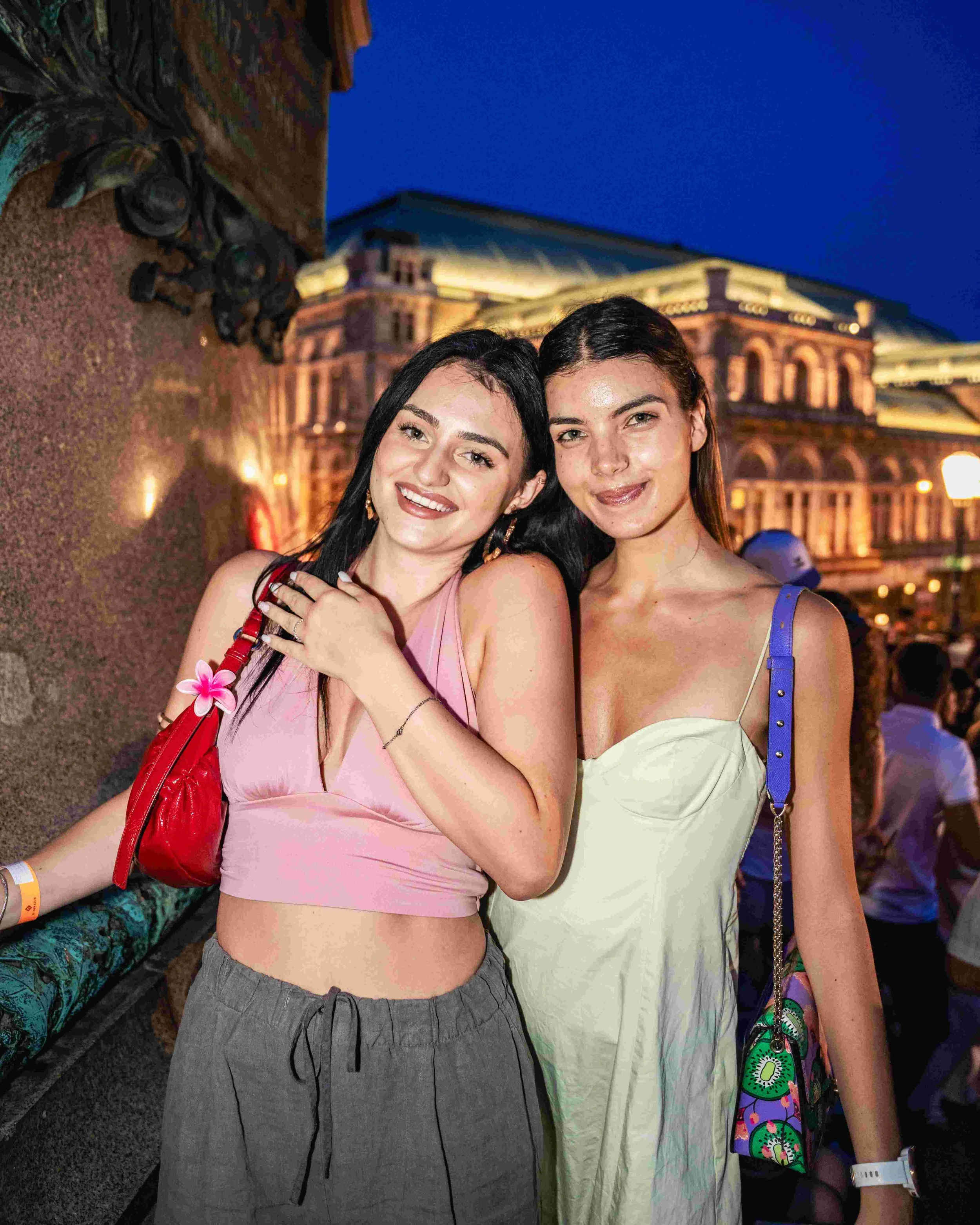 Two young women smiling and posing together at night, with an illuminated historic building in the background. One woman has long dark hair, wearing a pink top and gray pants, holding a red bag, with a pink flower hair accessory. The other woman has 