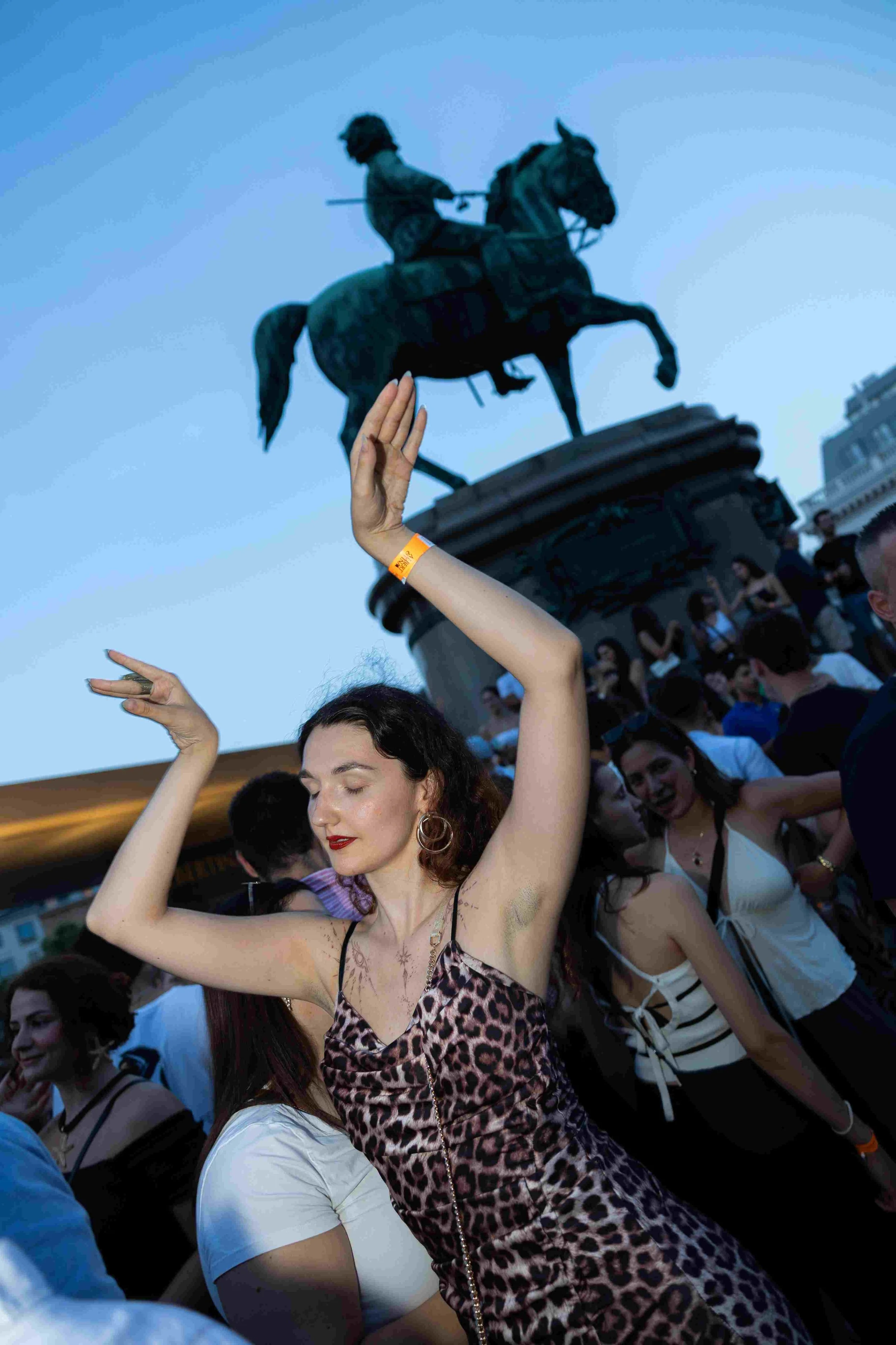 A woman dancing with her eyes closed and hands raised in front of a crowd at an outdoor event, with a statue of a mounted figure in the background.